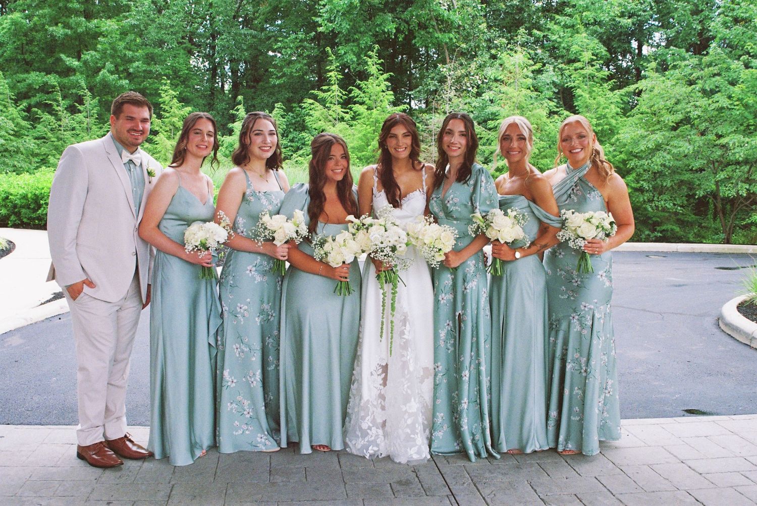 Bride in white lace gown poses with groom in tan suit and five bridesmaids in sage green dresses holding white bouquets.