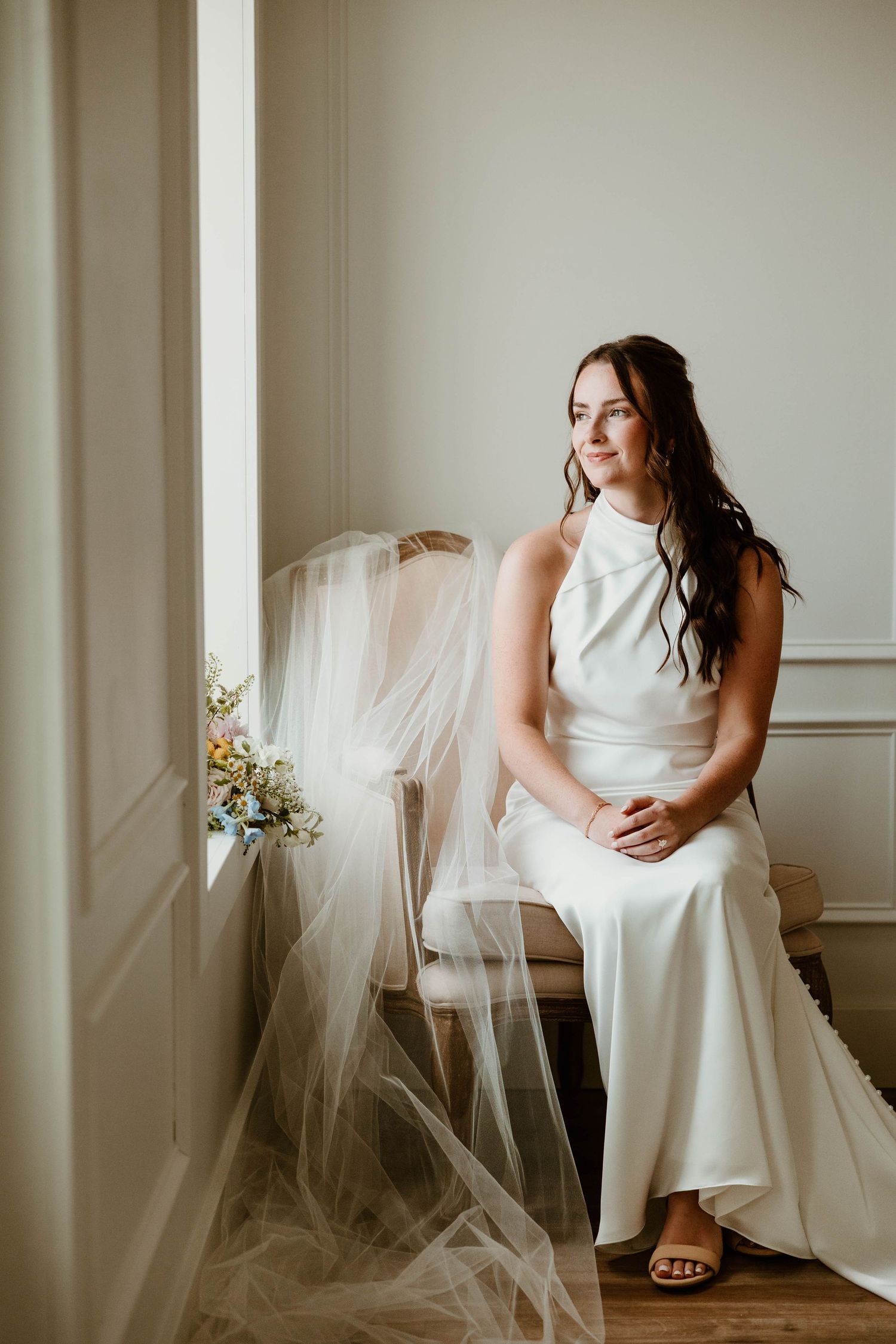 Bride in white halter gown sits by window with veil draped over chair and wildflower bouquet nearby.