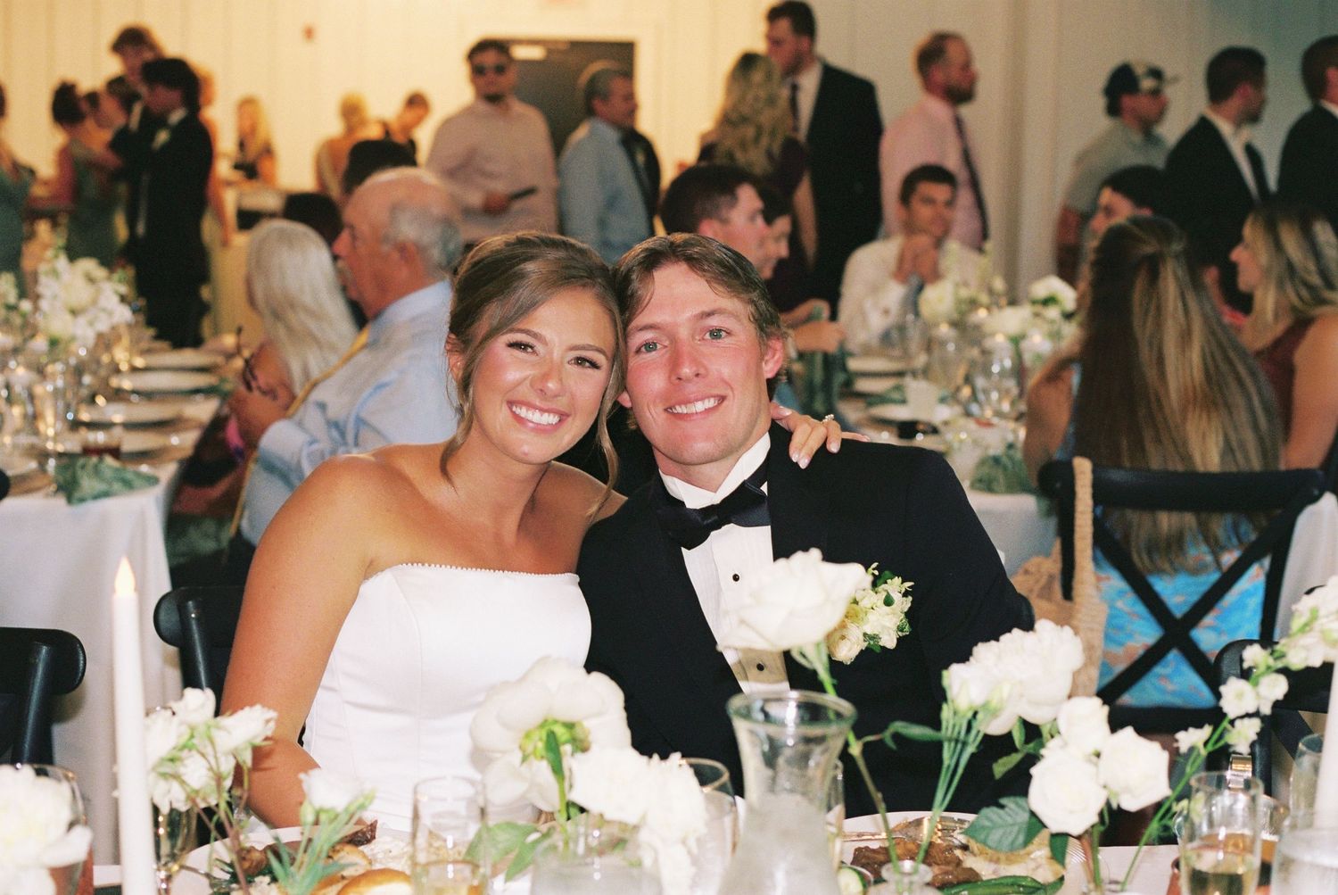 Bride and groom smiling at wedding reception table with white floral centerpieces and guests in background.