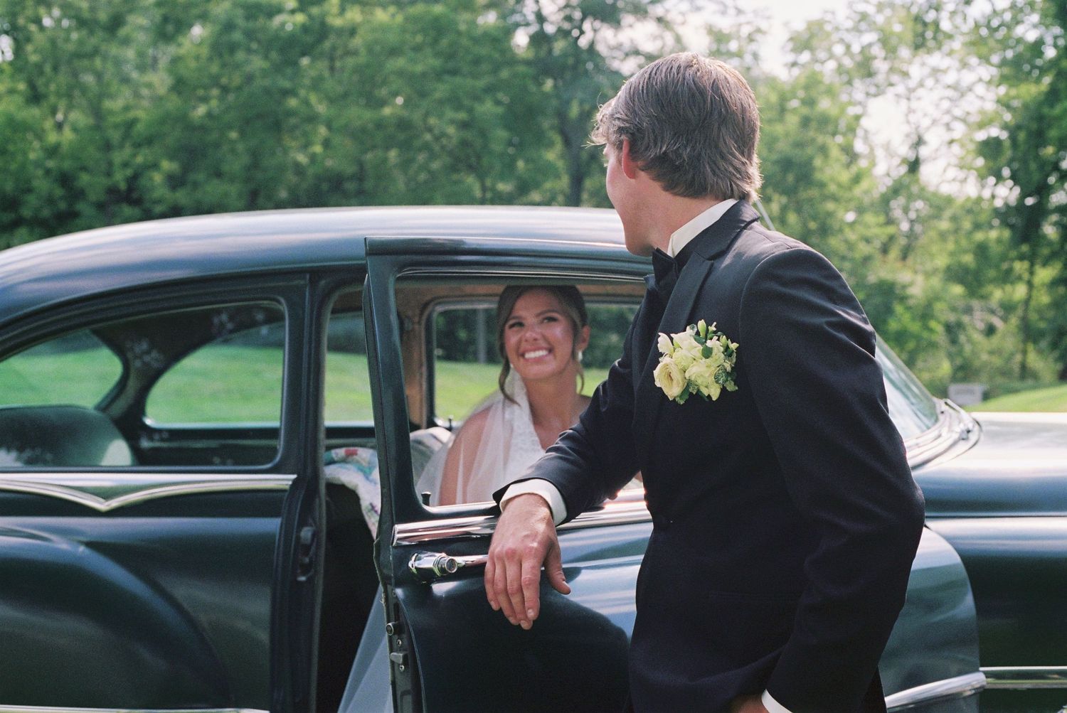 A smiling bride in a vintage black car is greeted by a groom in a tuxedo with a boutonniere at a wedding.