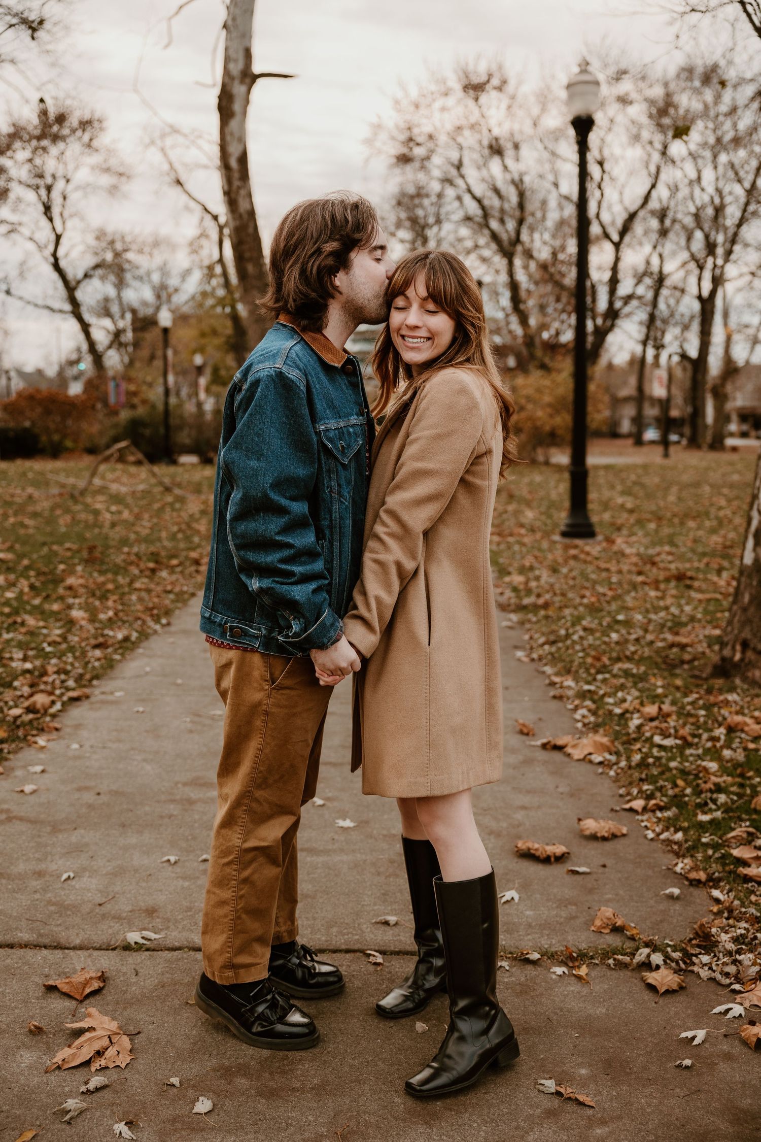 Stylish couple in autumn attire poses on leaf-covered path in park.