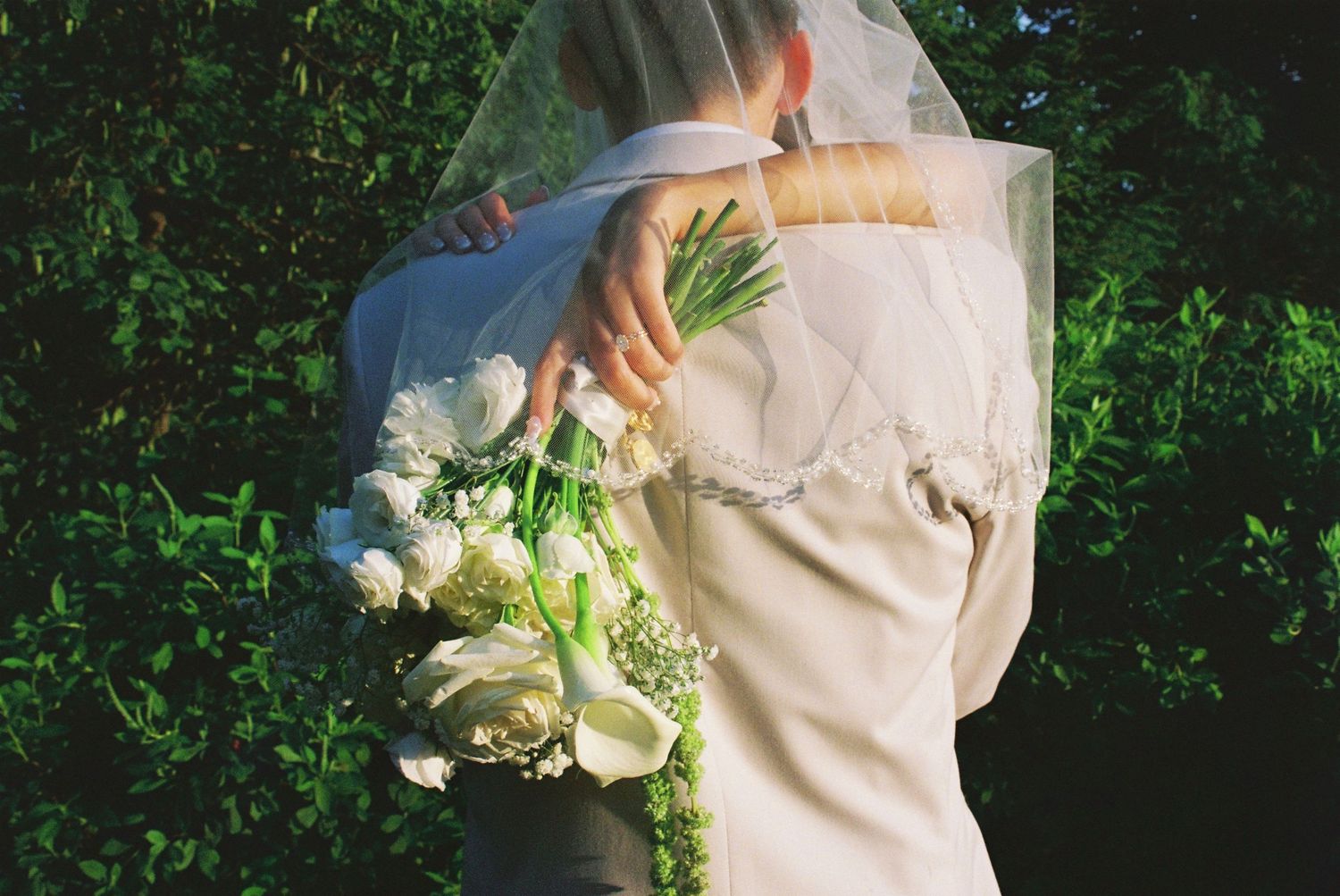 Bride and groom embracing outdoors, bride holding white calla lily bouquet with veil flowing, lush green foliage background.