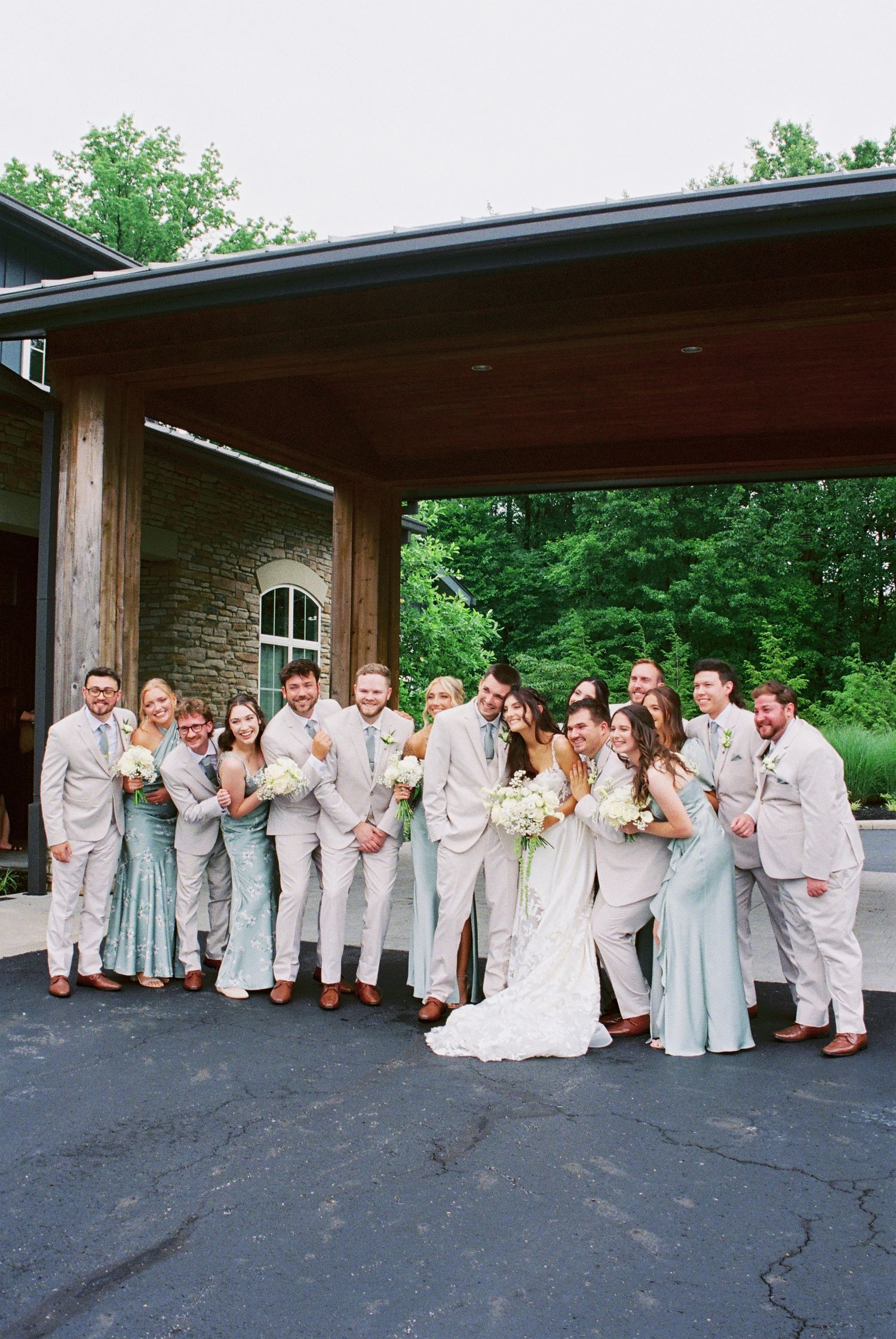 Wedding party poses together outside rustic stone venue, bridesmaids in sage green and groomsmen in tan suits.