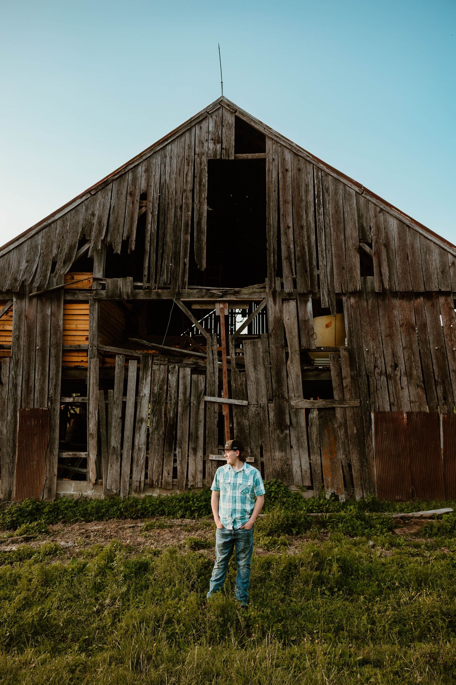 Person in blue shirt stands in front of weathered wooden barn in countryside setting.