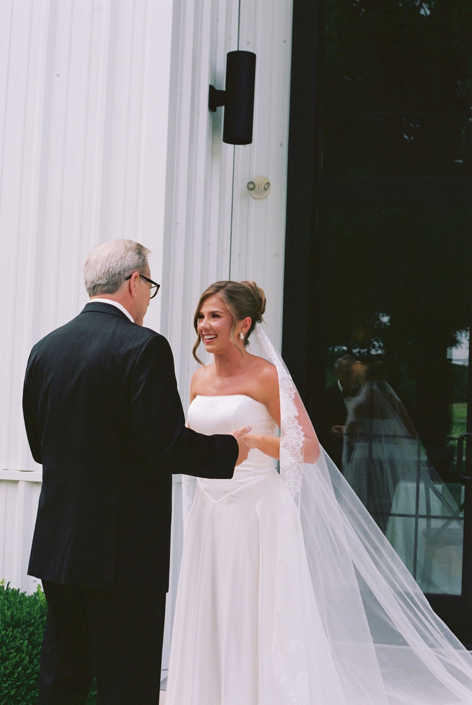 Bride in white gown and veil smiling at older man in black suit outside white building on wedding day.