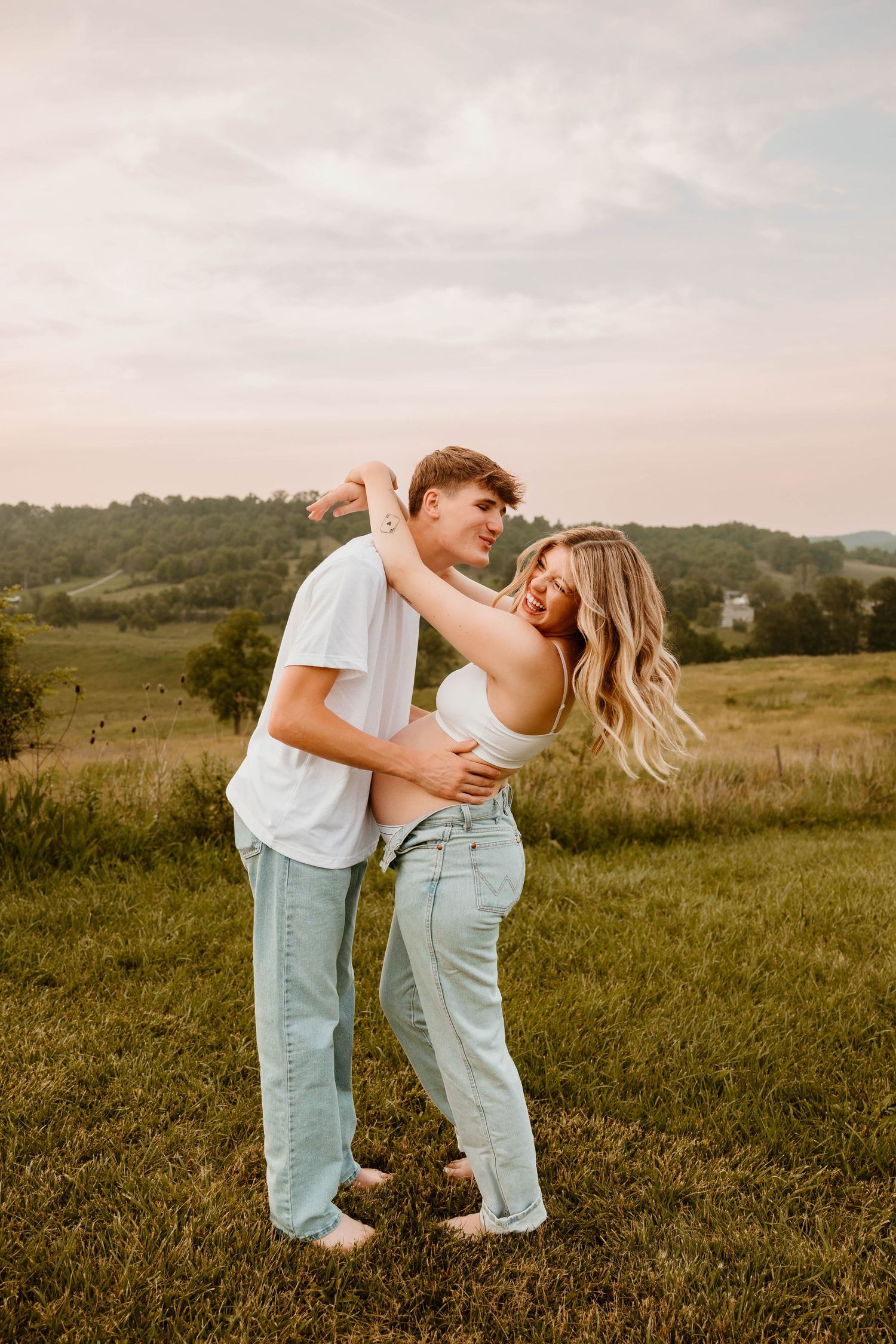A couple in matching white tops and light jeans embraces playfully in a grassy field at sunset.