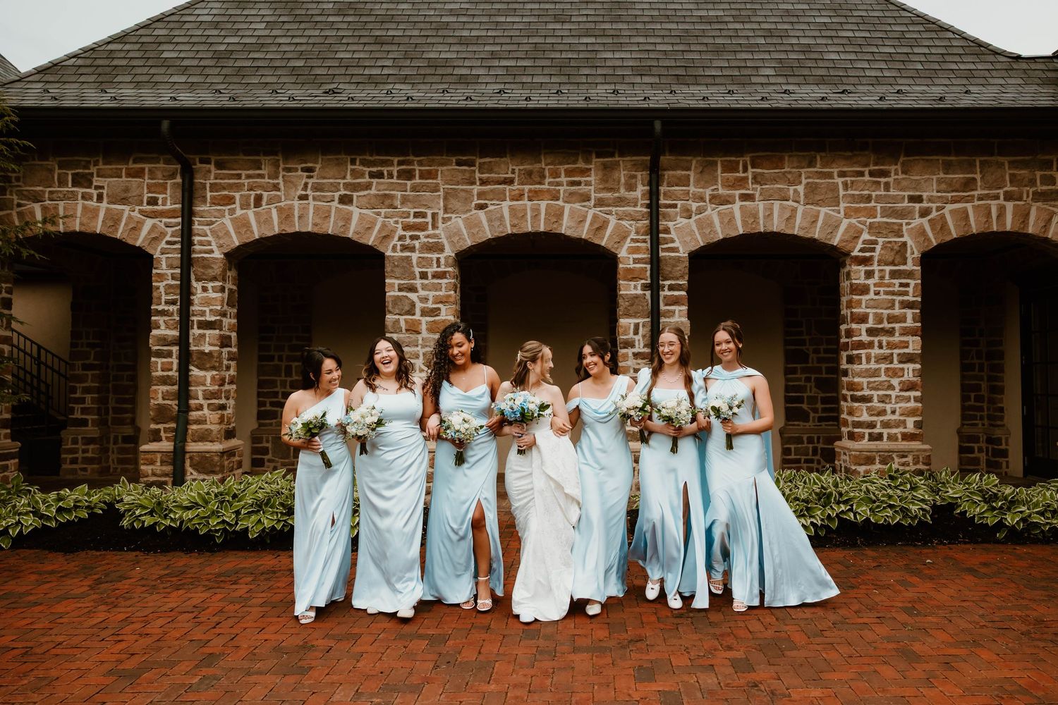 Bride and bridesmaids in light blue gowns holding bouquets pose in front of a stone building with arched windows.