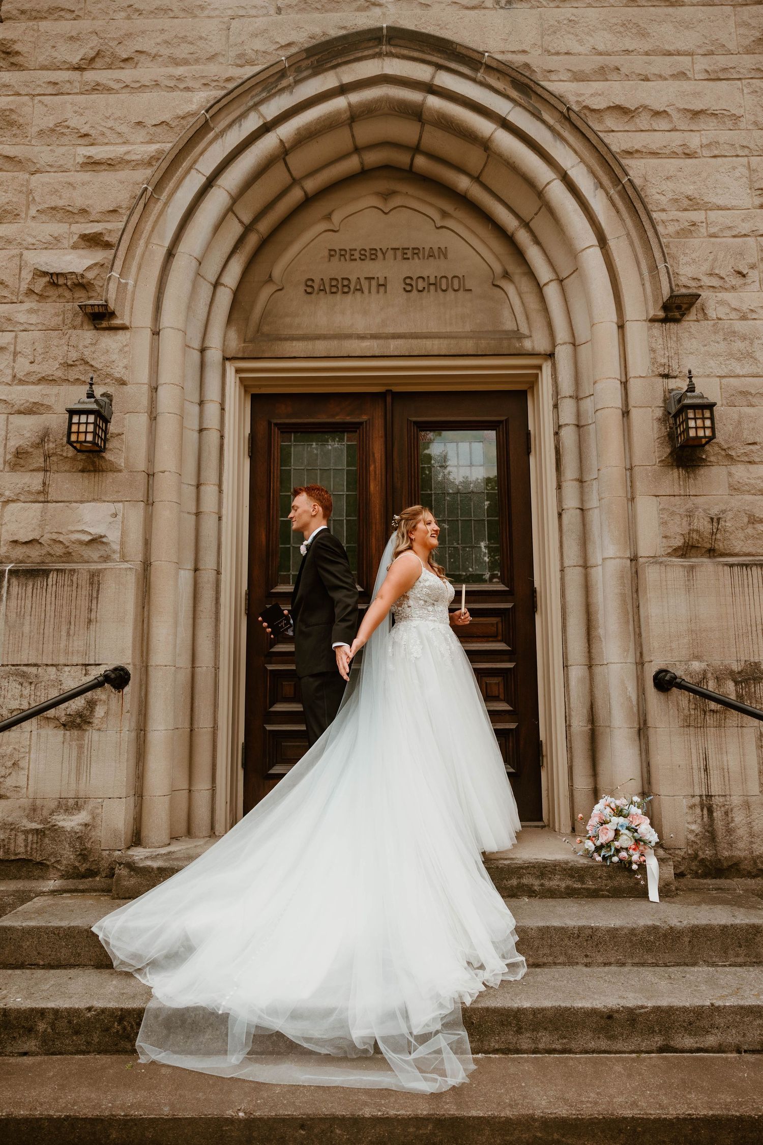 Bride and groom holding hands on stone church steps, her flowing white gown billowing, beside a Presbyterian Sabbath School entrance.