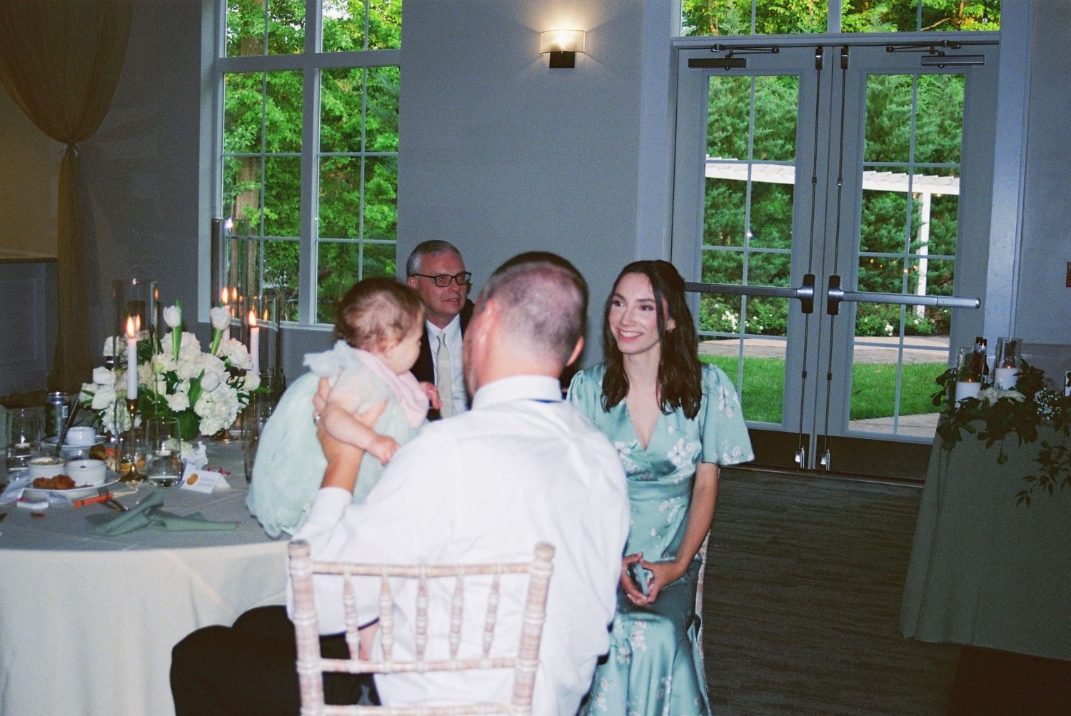 Guests in elegant attire gather around a decorated reception table in a bright, garden-view wedding venue.
