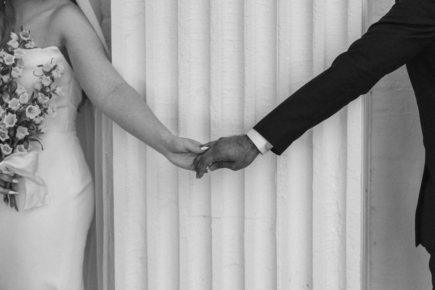 A black and white photo of newlyweds holding hands with formal wedding attire.