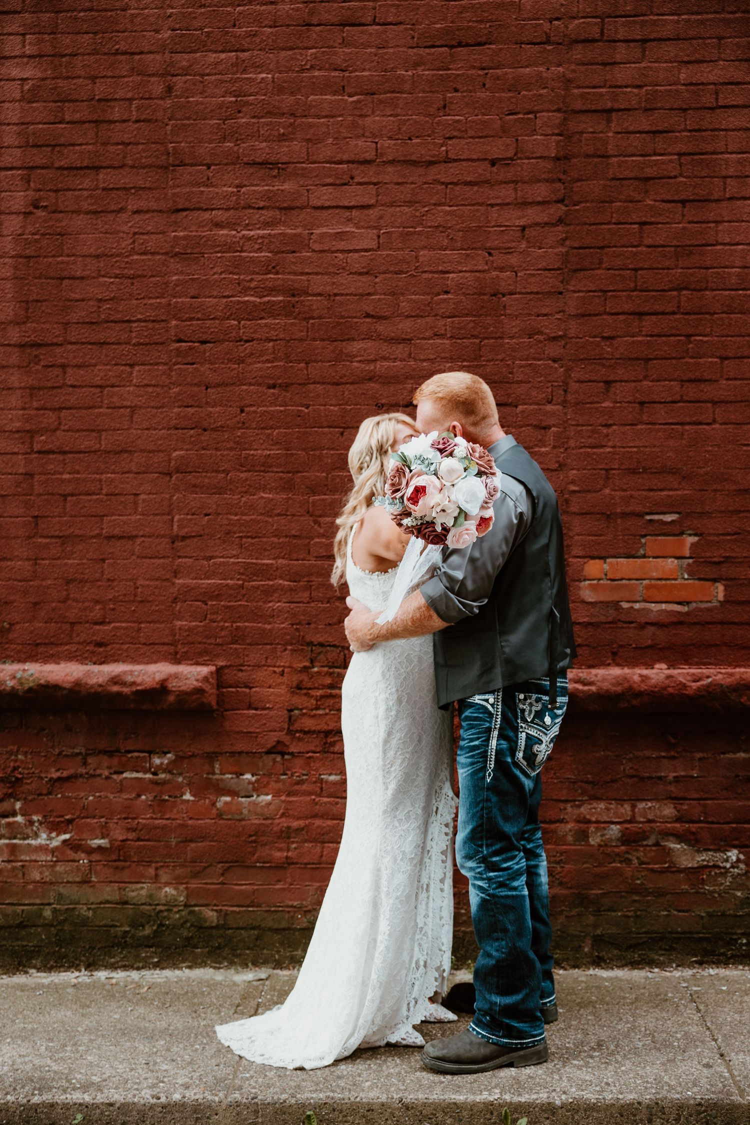 A couple shares an intimate moment against a rustic red brick wall on their wedding day.