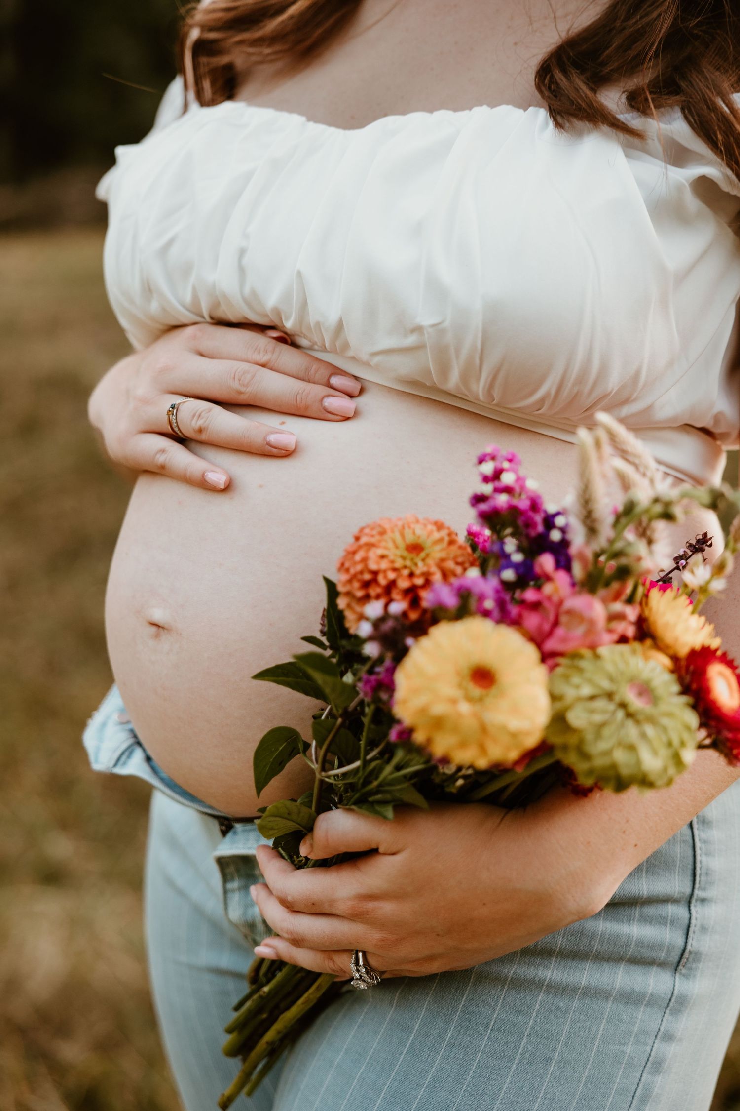 A pregnant woman in light clothing holds a colorful bouquet of flowers against her belly in an outdoor setting.