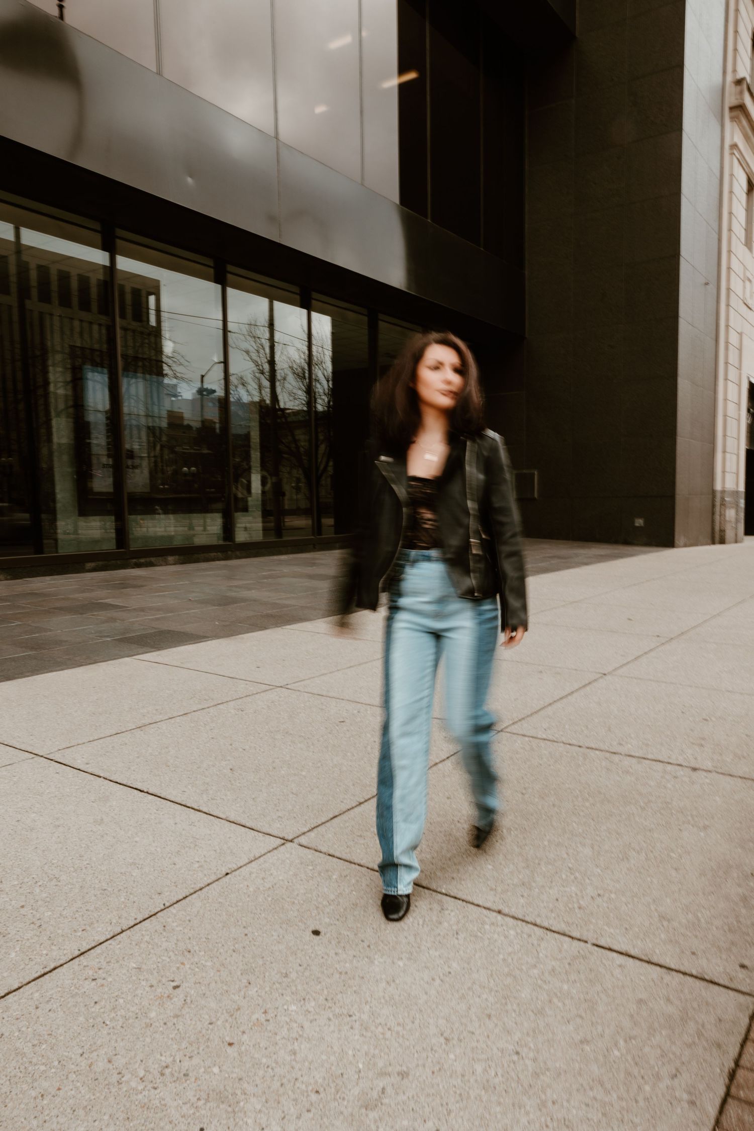 Woman in leather jacket and jeans walks confidently on urban sidewalk.
