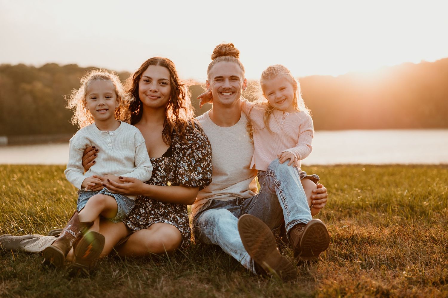 Family sitting together on grass during golden hour sunset by a lake in matching outfits.