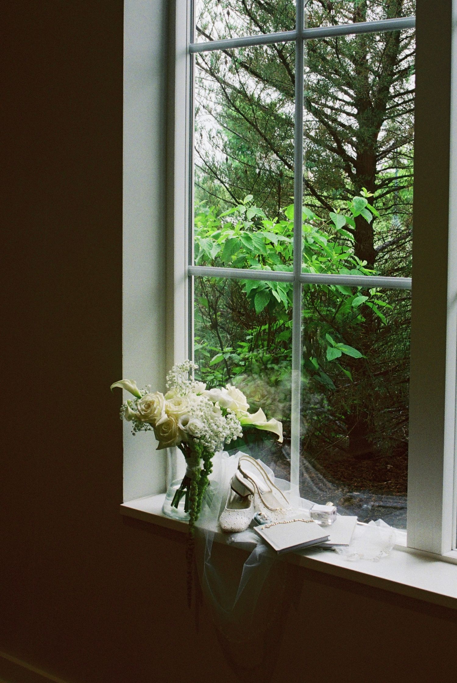 Bridal bouquet of white flowers and wedding shoes on a windowsill overlooking lush green trees.