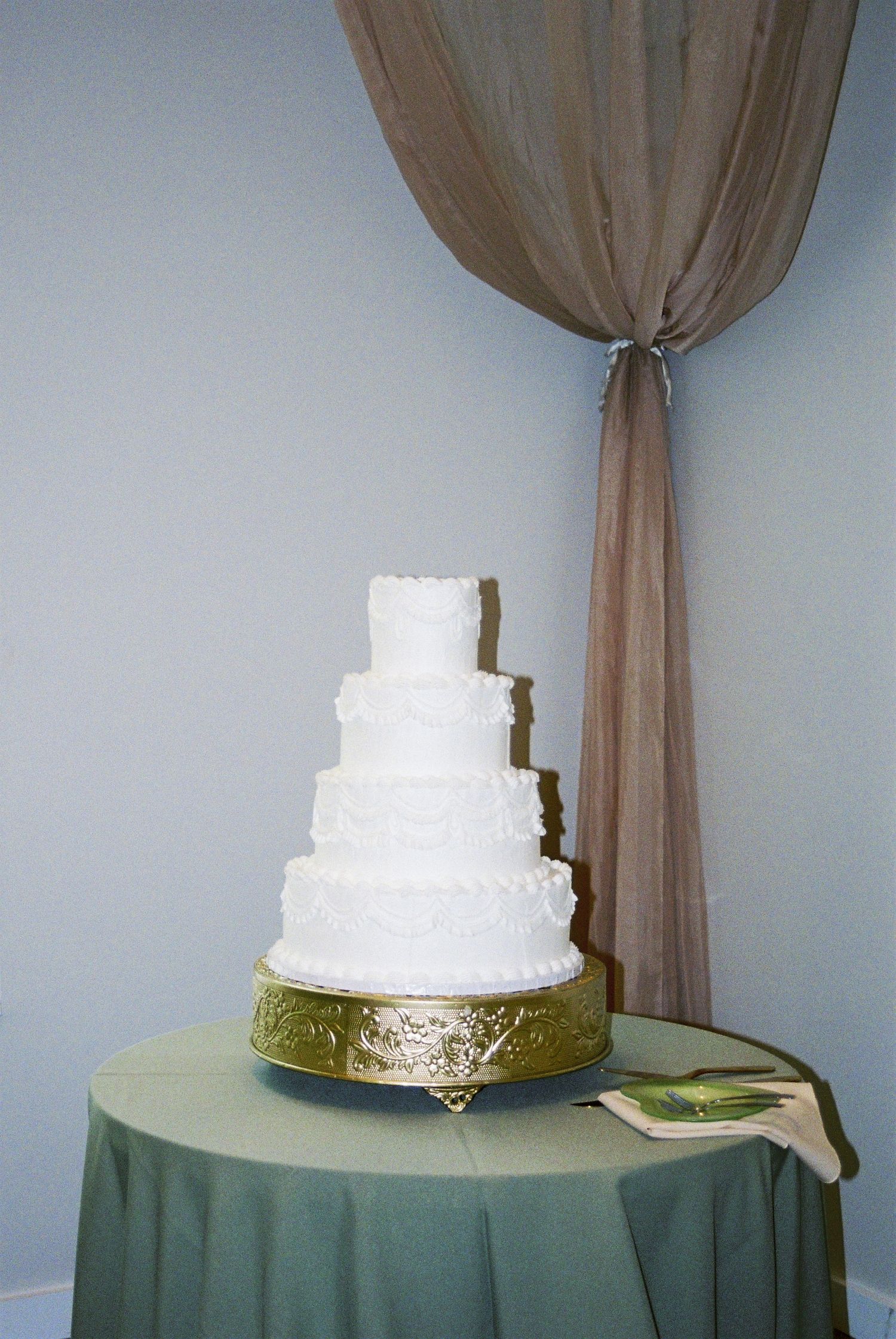 Elegant four-tier white wedding cake on gold stand displayed on sage green tablecloth with draped curtain.