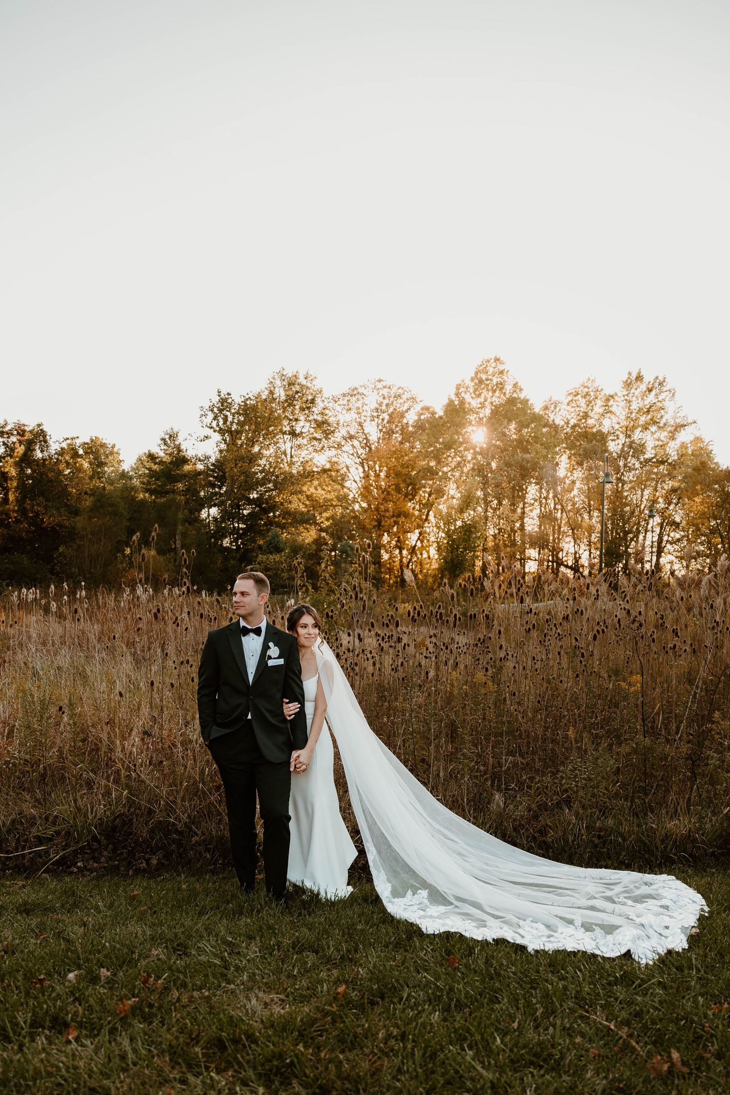 Wedding couple poses in field at sunset with dramatic long cathedral veil trailing behind.