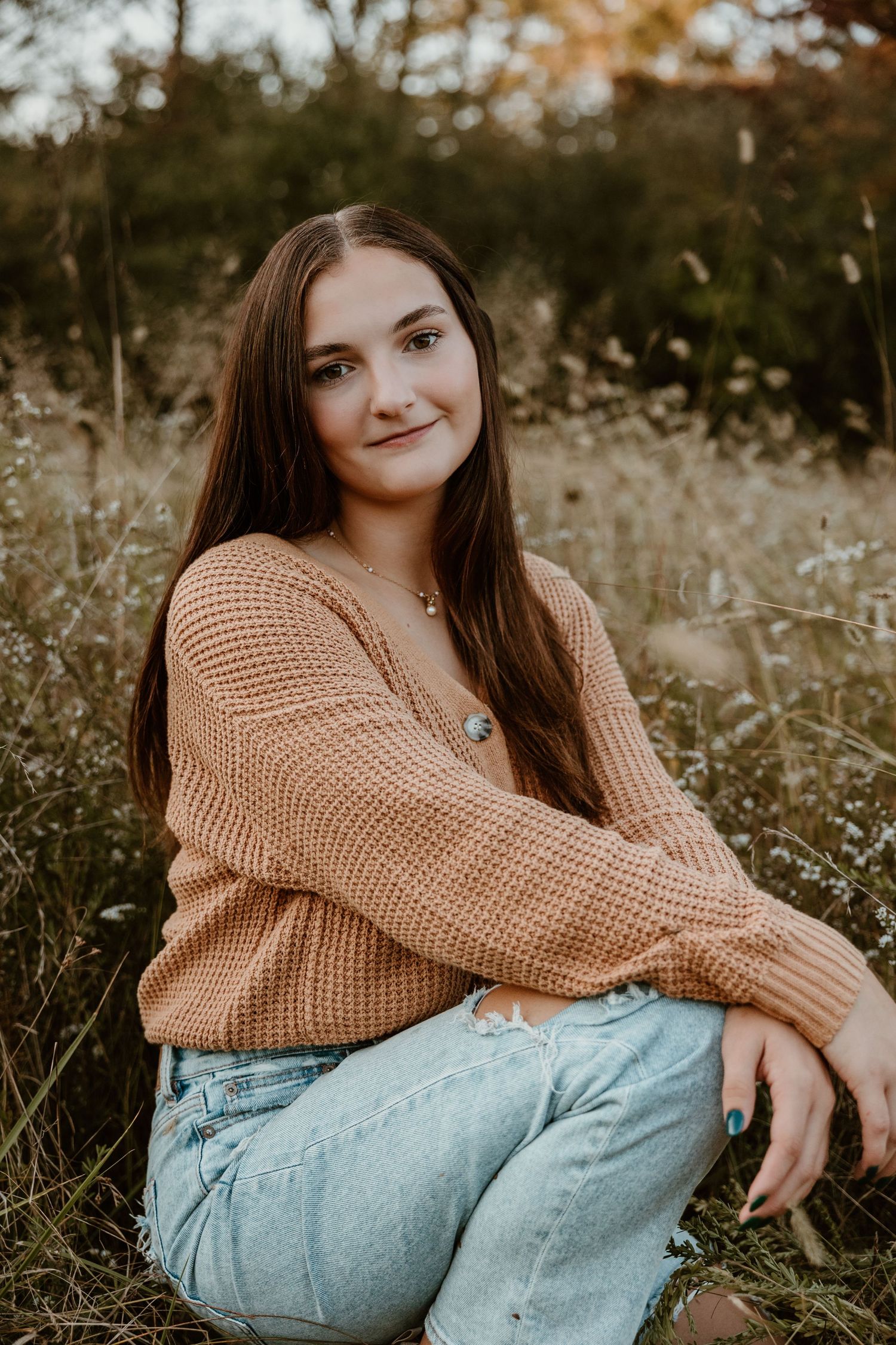 Young woman in tan sweater and jeans poses in autumn field with wildflowers.