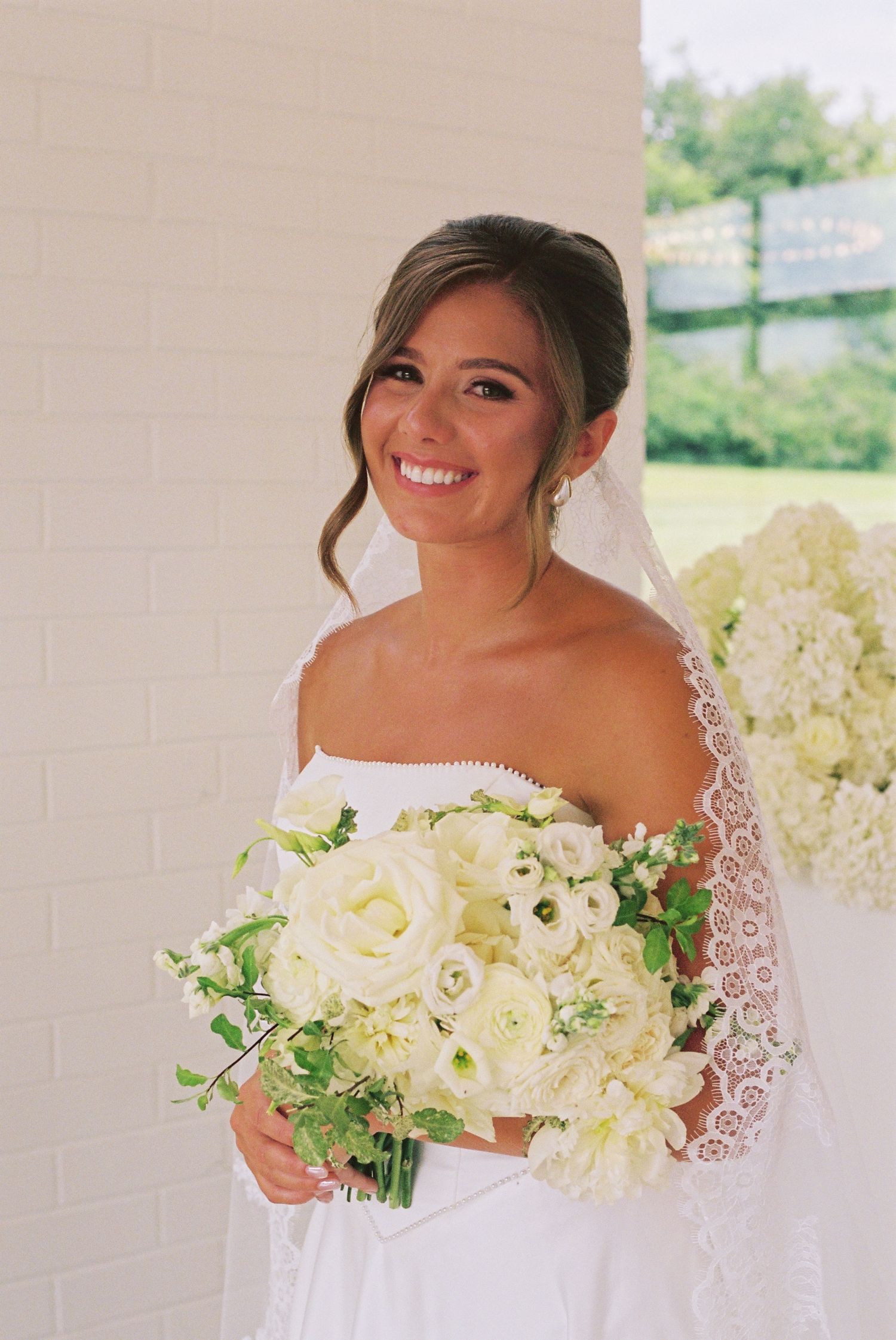Smiling bride in white off-shoulder gown holds lush white rose bouquet, wearing lace veil, posing near brick wall.