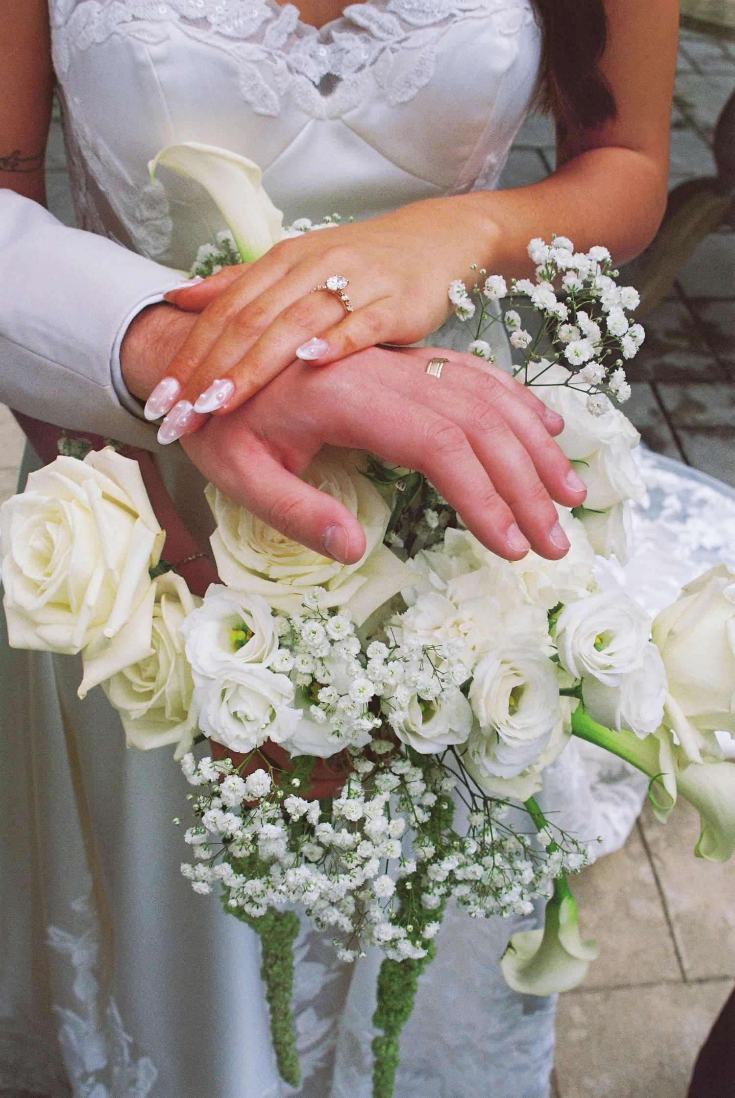 Bride and groom displaying wedding rings on hands resting over a bouquet of white roses and baby's breath.