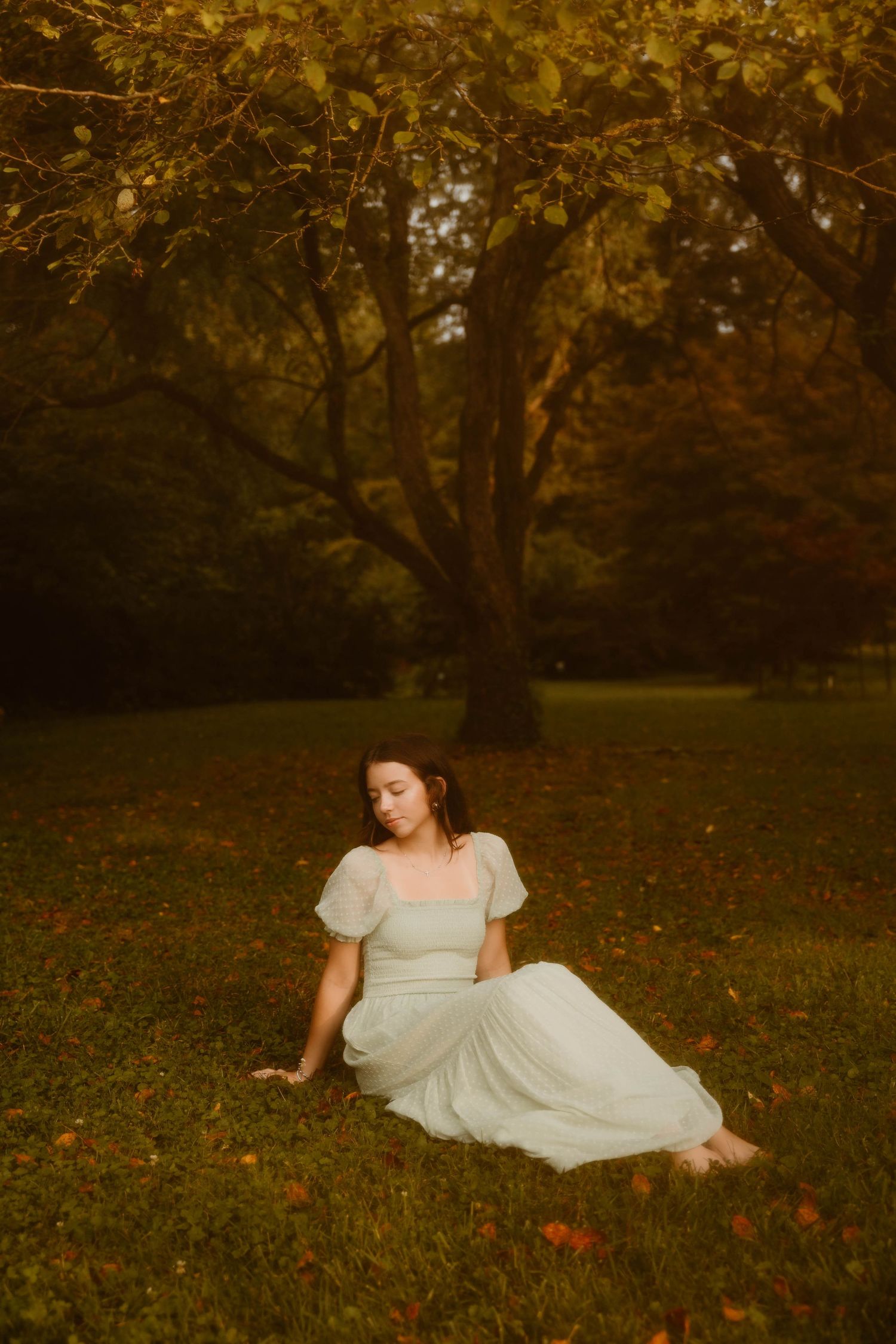 A dreamy portrait of someone in a white dress resting on autumn grass under a golden-lit tree.