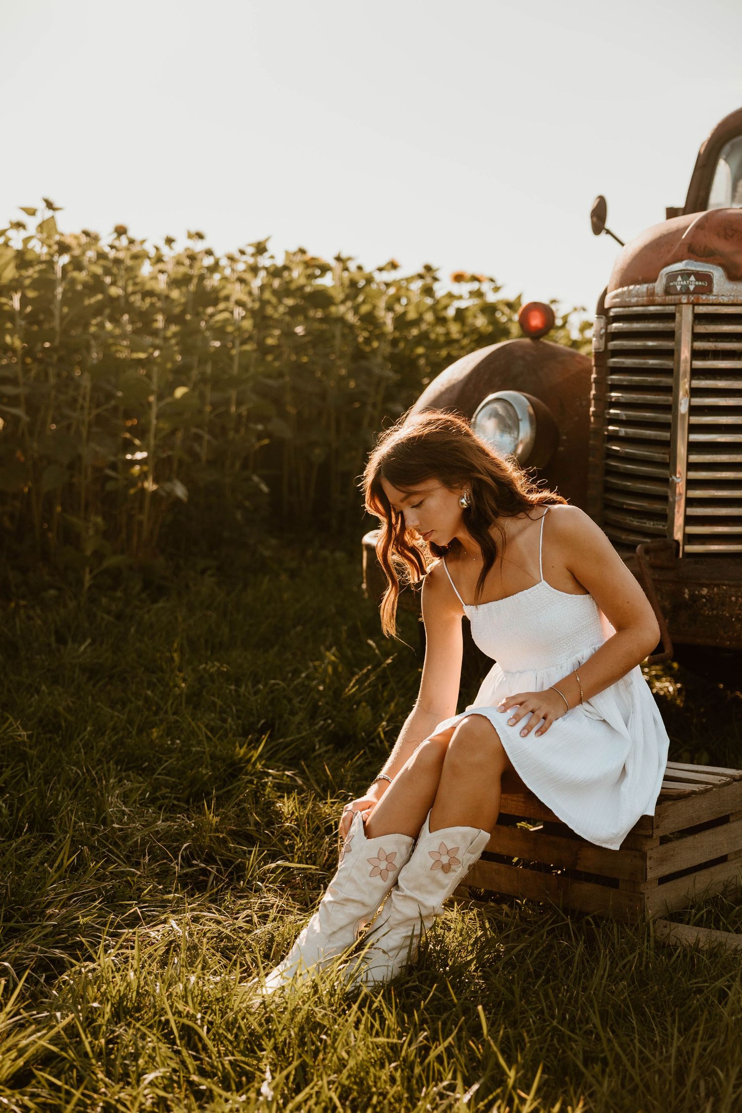 A person in a white dress sits thoughtfully beside a vintage tractor in golden sunlight.