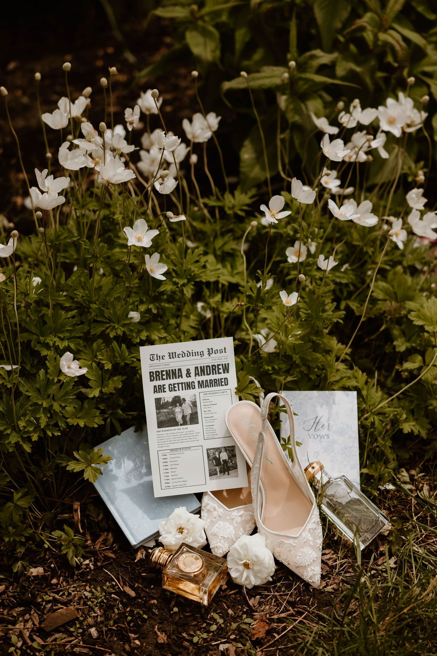 Bridal flat lay with lace heels, perfume, and wedding newspaper announcement among white anemone flowers.