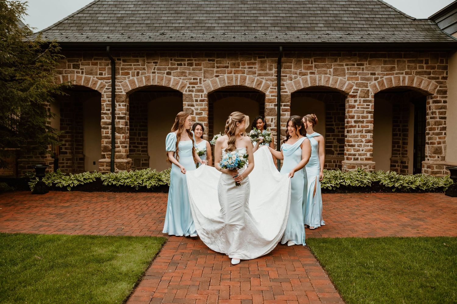 Bridesmaids in light blue gowns surround a bride in a white dress outside a stone building on a brick pathway.