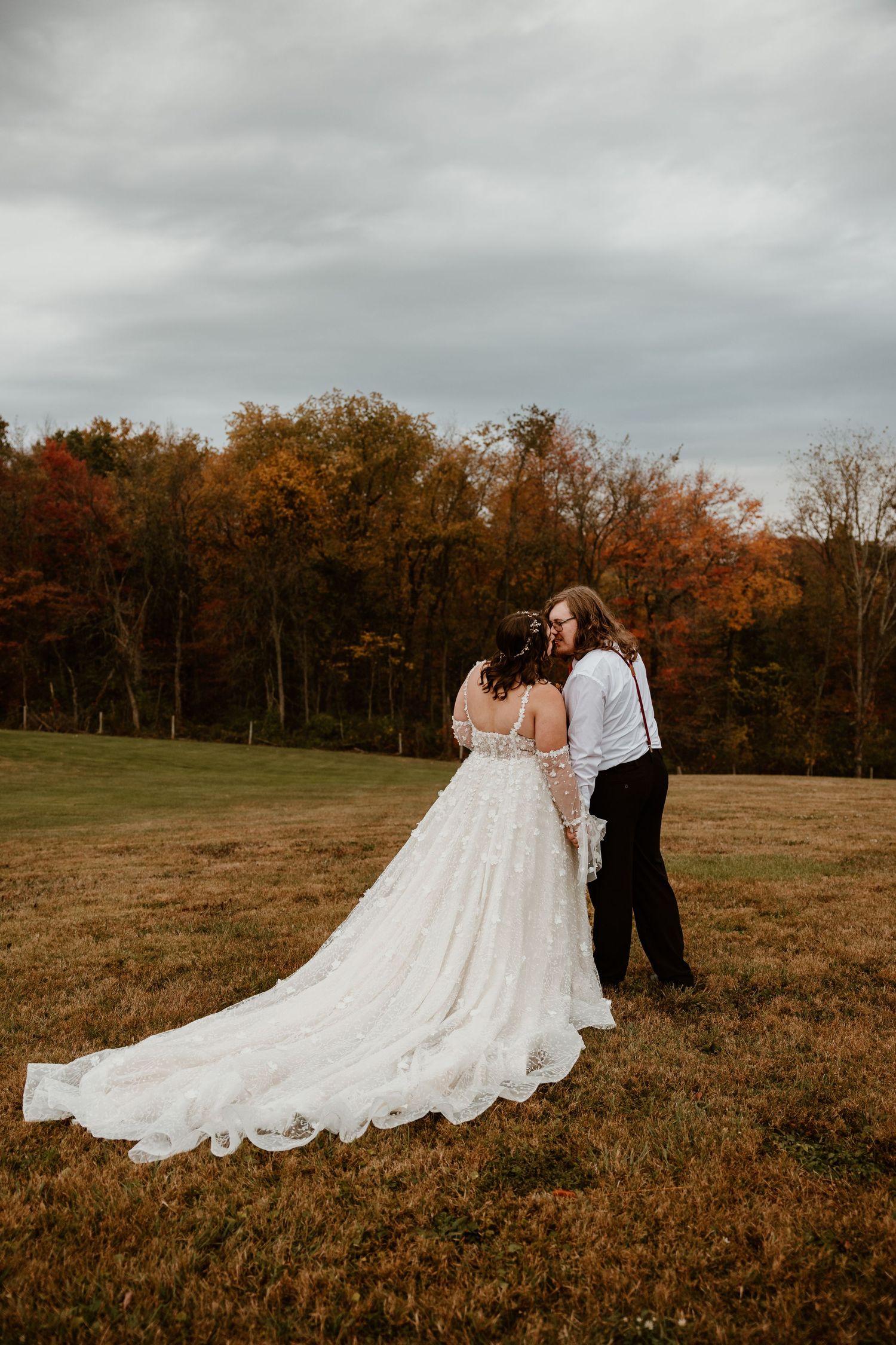 Couple in wedding attire poses in autumn field with dramatic cloudy sky.