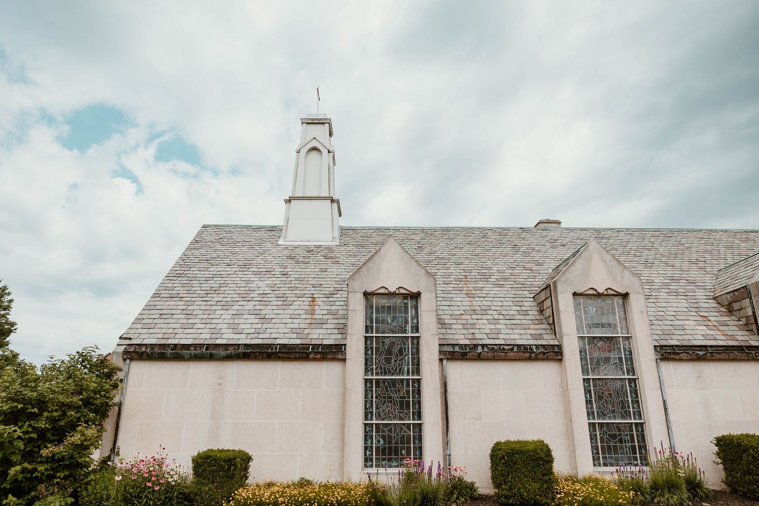 Historic white stone church with gray slate roof, Gothic windows, and white steeple against cloudy sky.