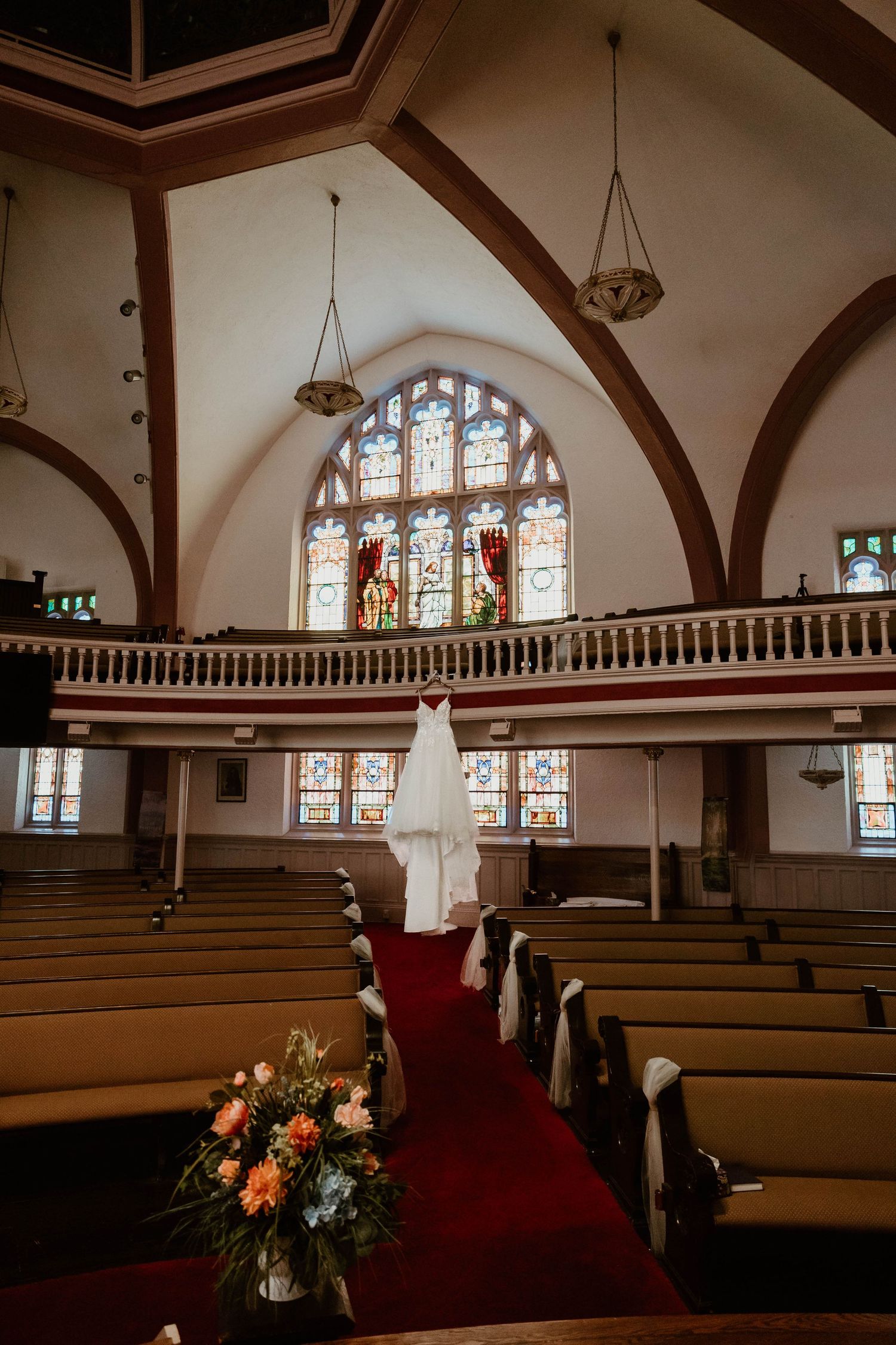 Wedding dress hanging in a church aisle with stained glass windows, red carpet, wooden pews, and floral arrangement.