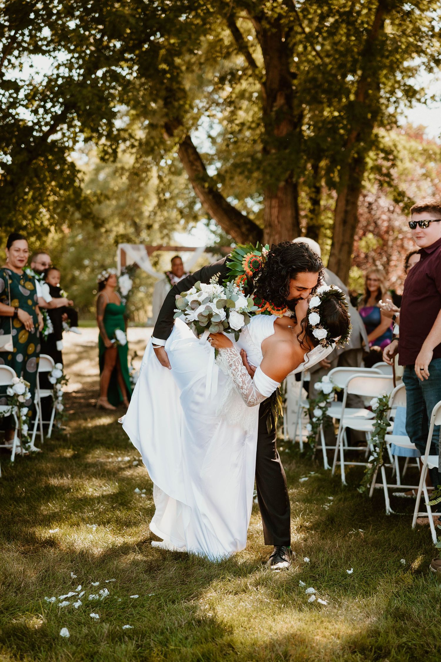 Joyful outdoor wedding celebration with guests seated on white chairs under trees.