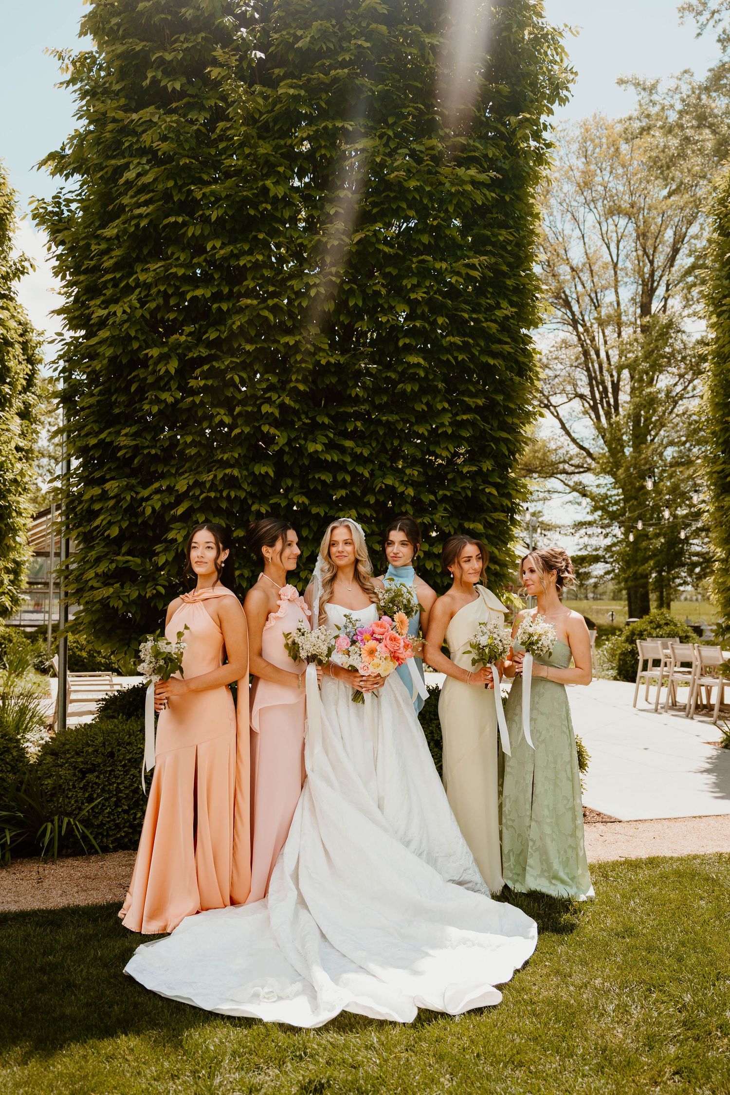 Bride in white gown with bridesmaids in peach and sage green dresses holding floral bouquets outdoors.