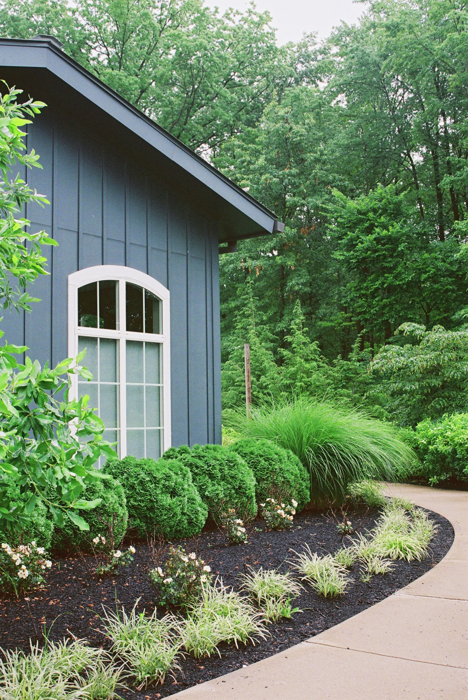 Blue barn-style building with arched window surrounded by lush green landscaping and manicured shrubs.