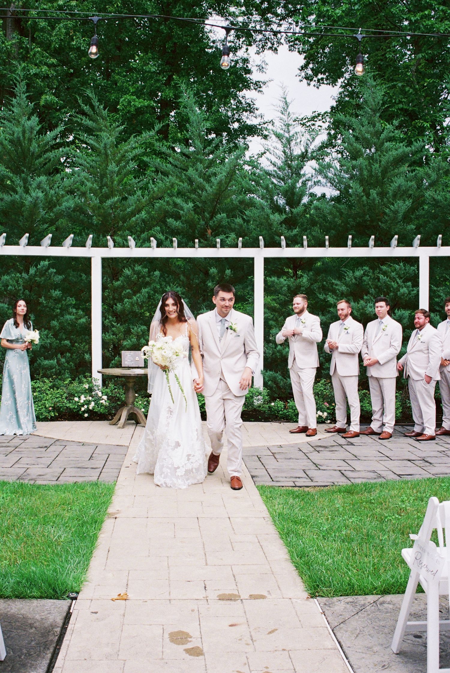 Newlyweds walk down the aisle at outdoor ceremony with wedding party standing beneath white pergola.