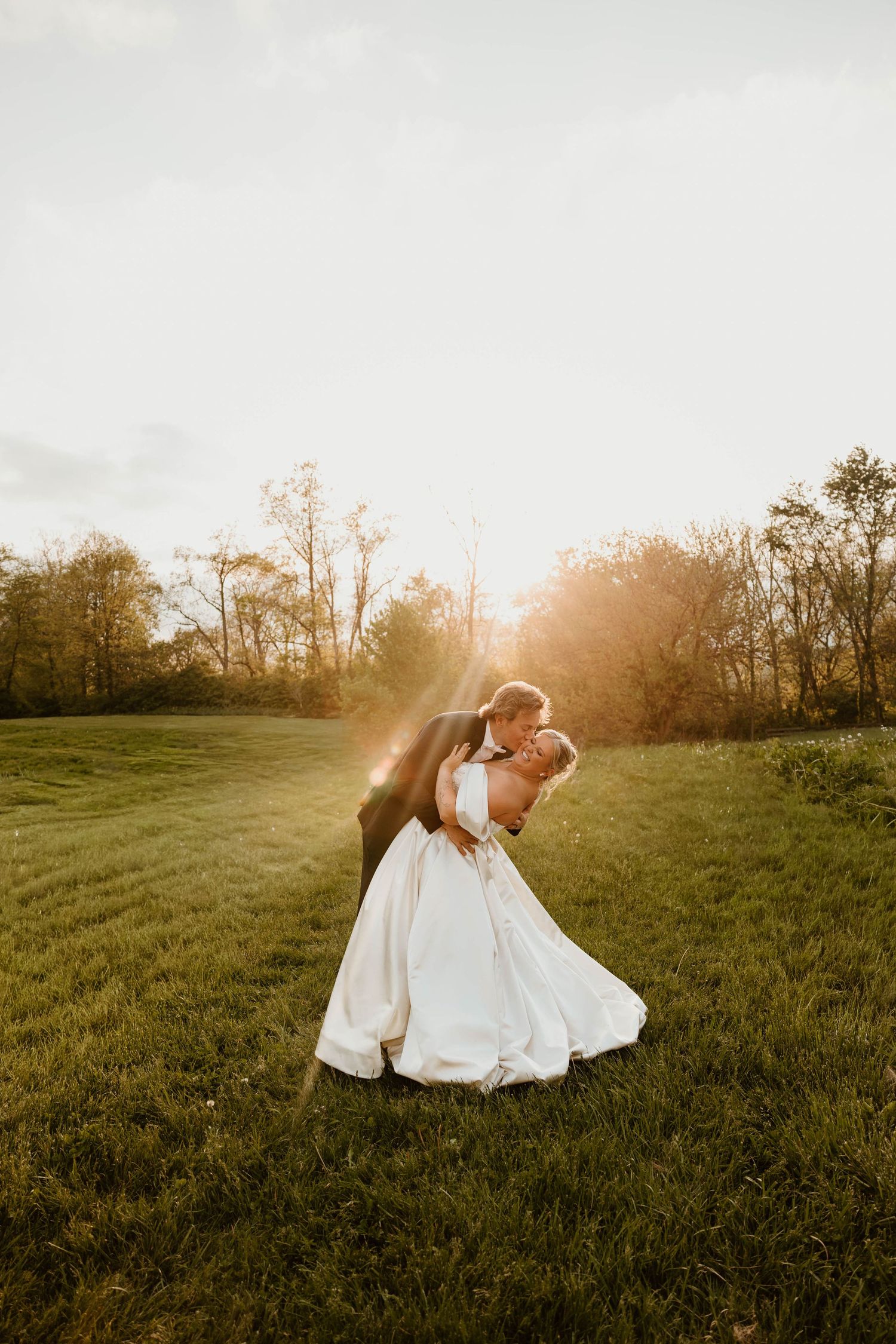 Bride and groom share a romantic dip kiss at golden sunset in a lush green field surrounded by trees.