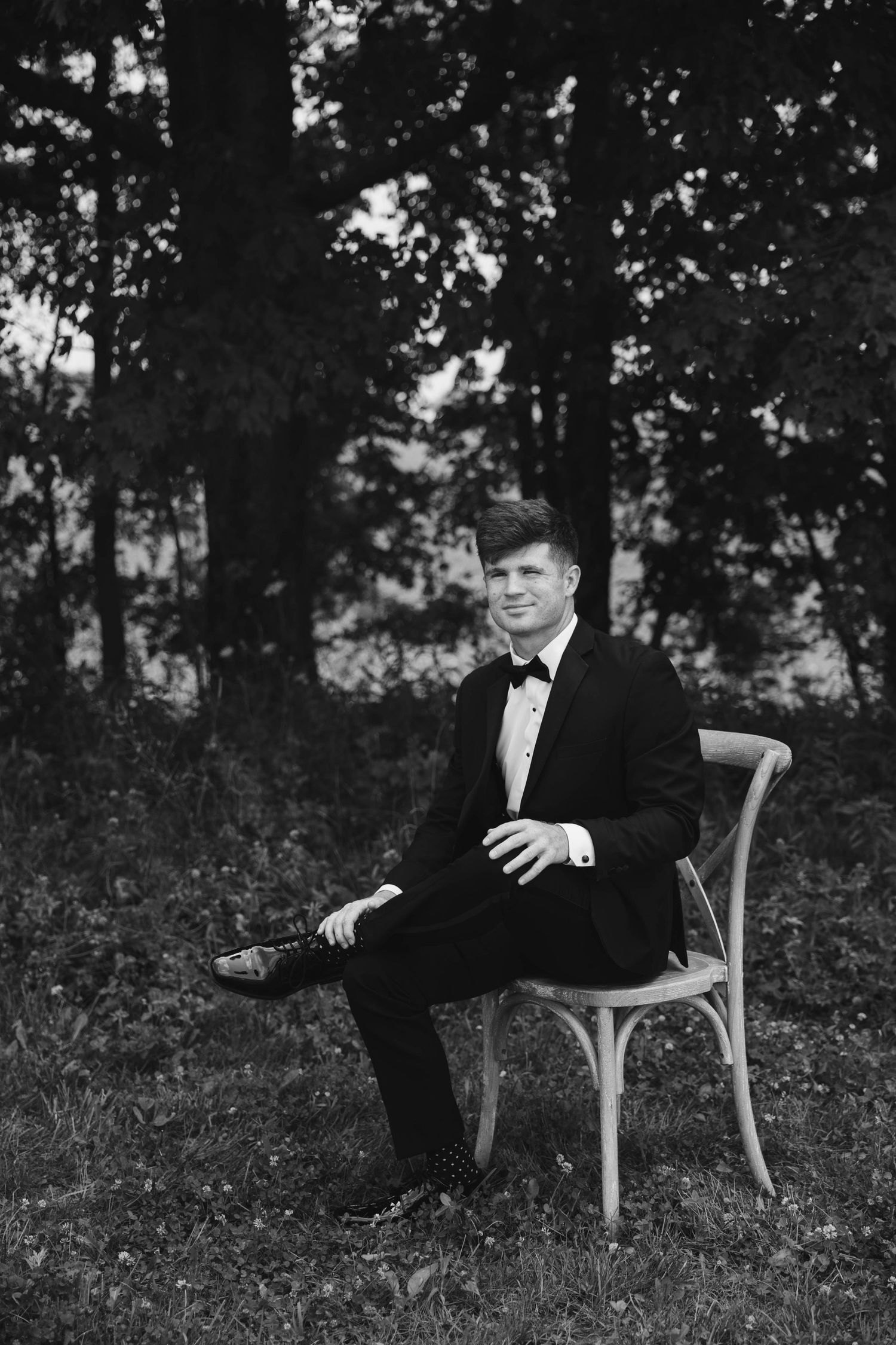 A young man in a tuxedo sits on a wooden chair outdoors, posing confidently among trees in a black and white photo.