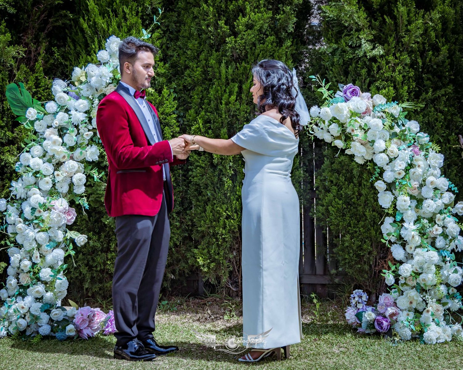 A couple in red and white attire exchanges rings beneath a floral arch during their garden ceremony.