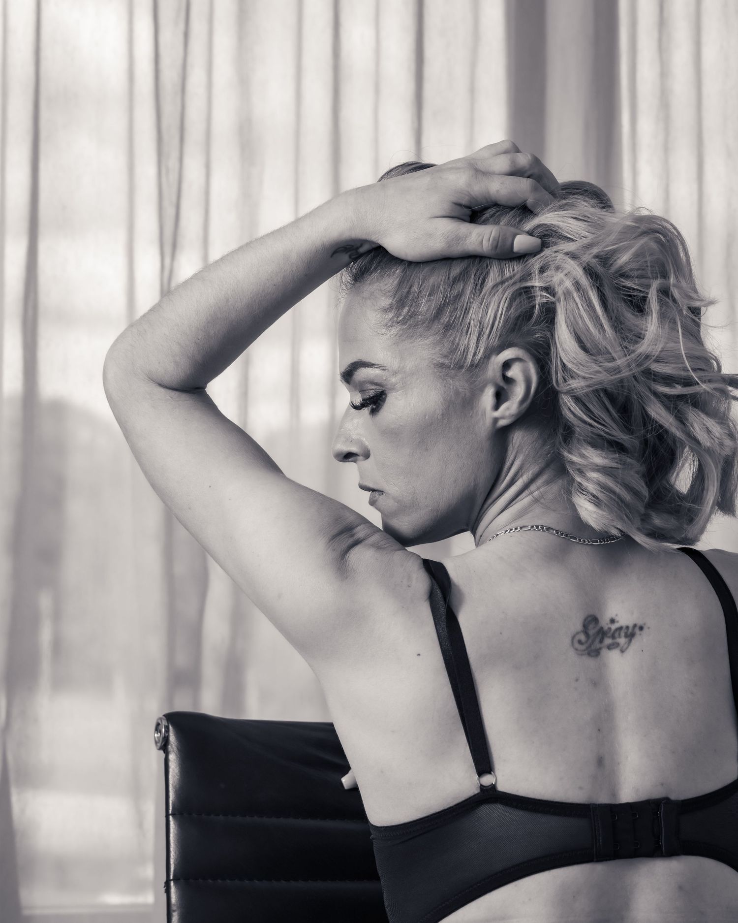 Black and white artistic portrait showing back tattoo and blonde styled hair against soft window light.