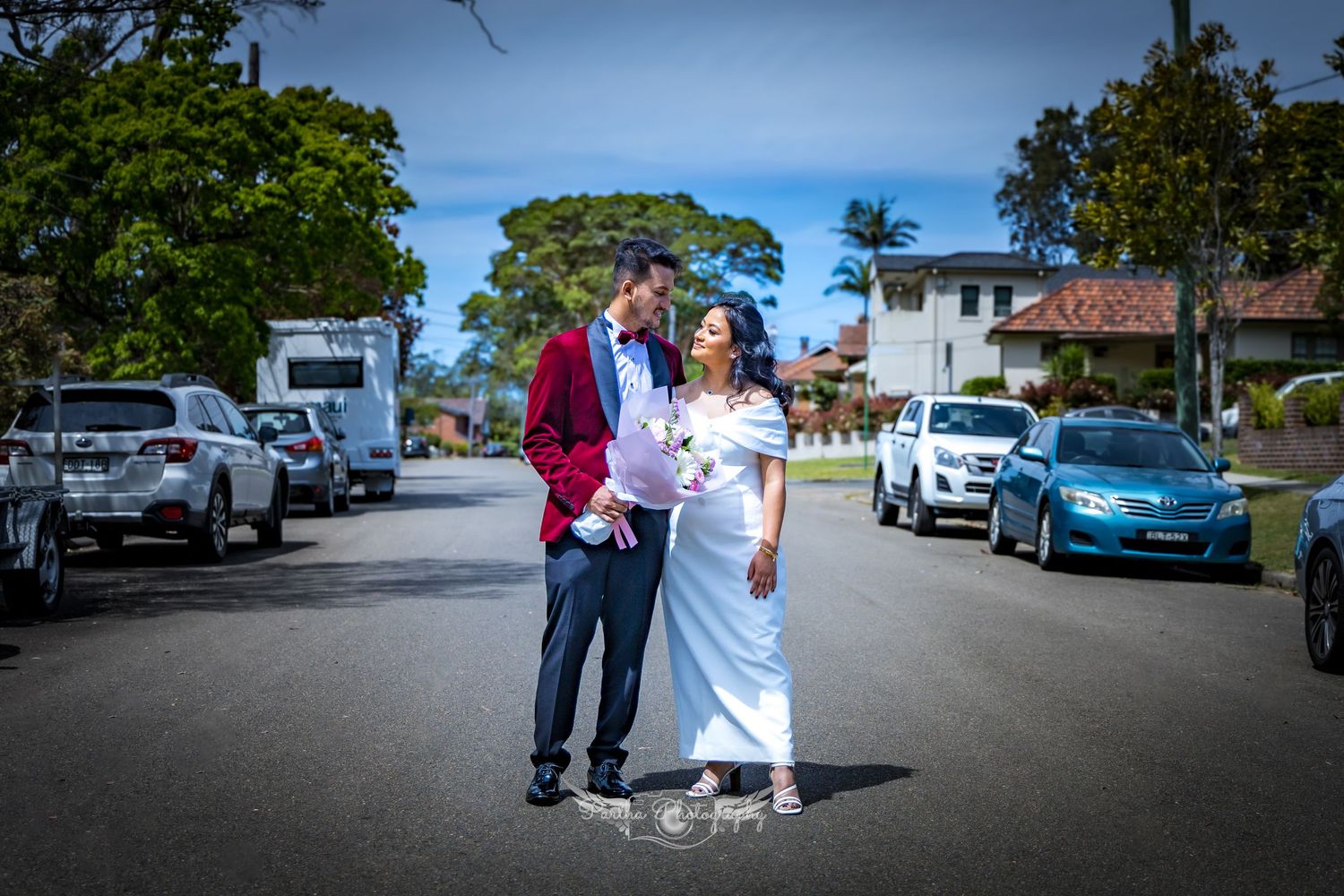 Newlyweds walking together on a street lined with parked cars.
