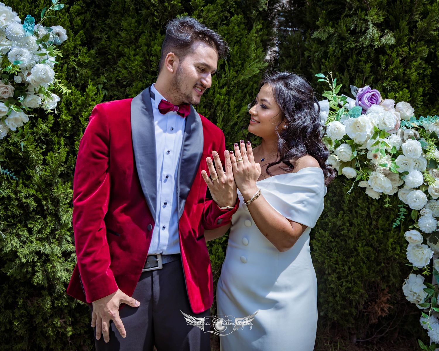 A couple in red and white wedding attire sharing a joyful moment under a floral arch.