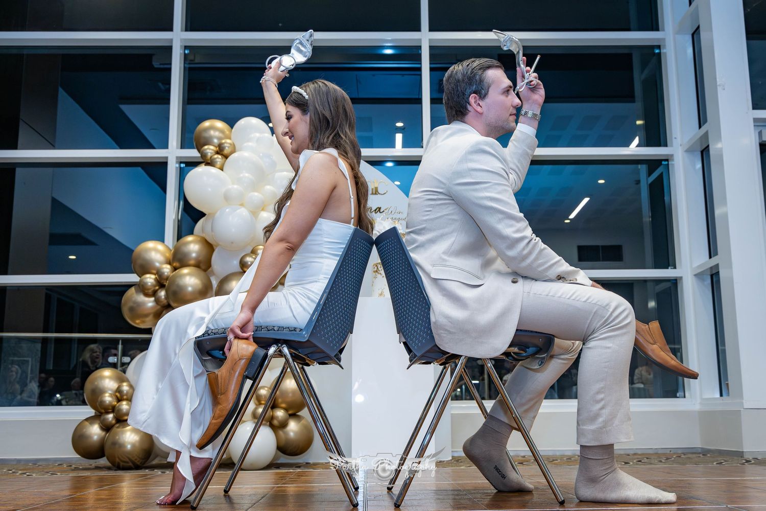 Playful couple poses back-to-back on chairs at wedding reception with gold balloon arch.