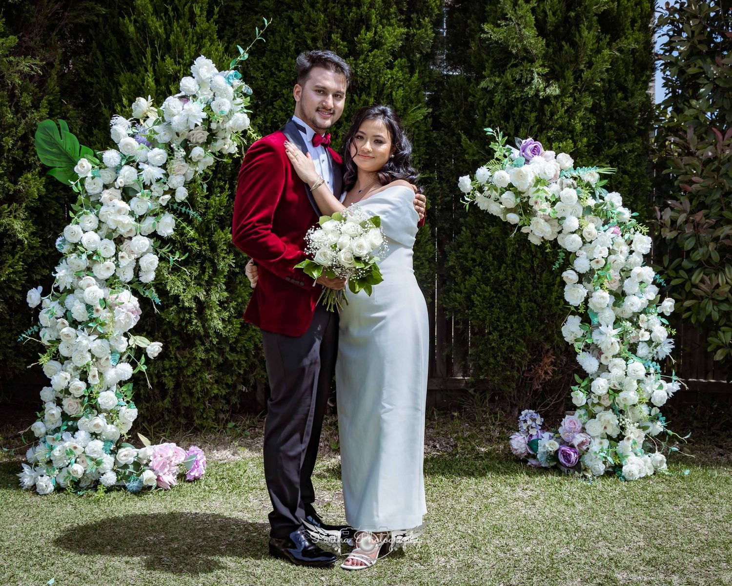 Couple poses under elegant white floral arch during outdoor garden wedding ceremony.