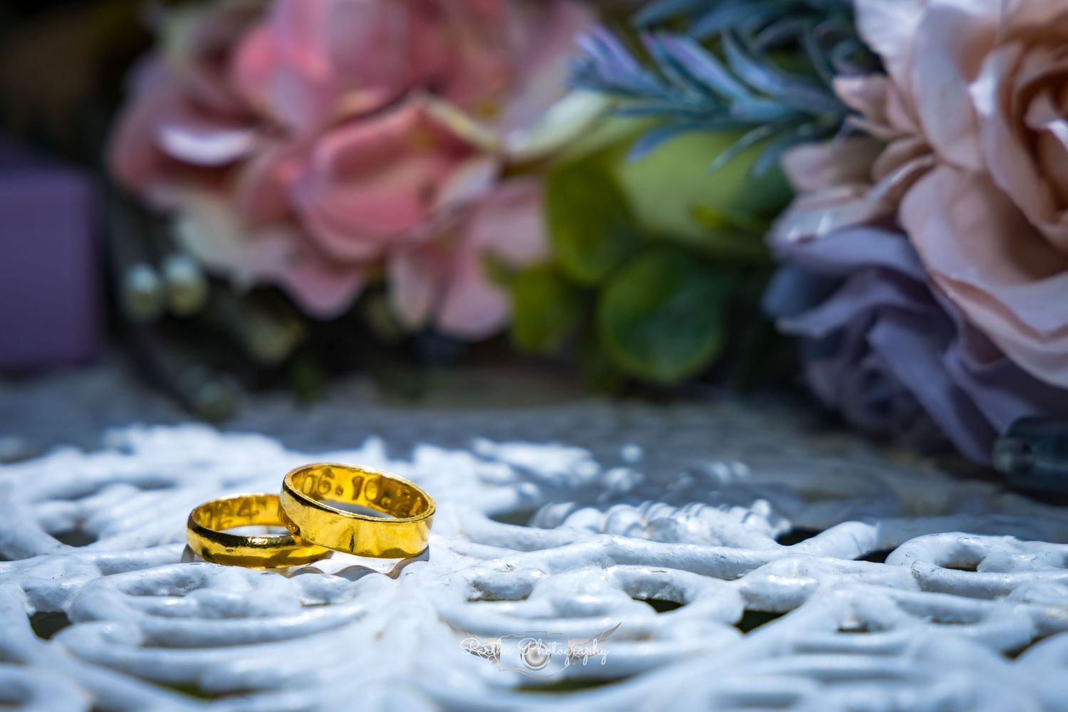 Two golden wedding rings displayed on white lace with soft pastel floral decorations in the background.