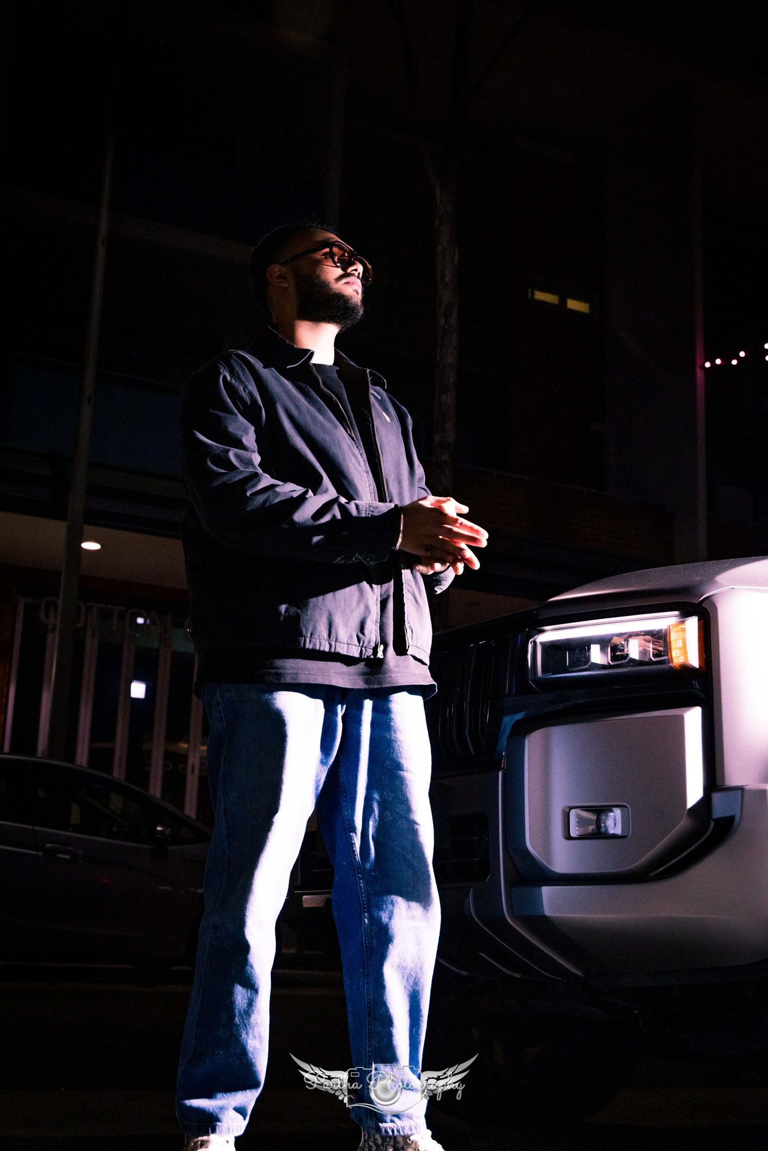 A person in baggy jeans and dark jacket stands confidently in dramatic nighttime lighting next to a vehicle corner.