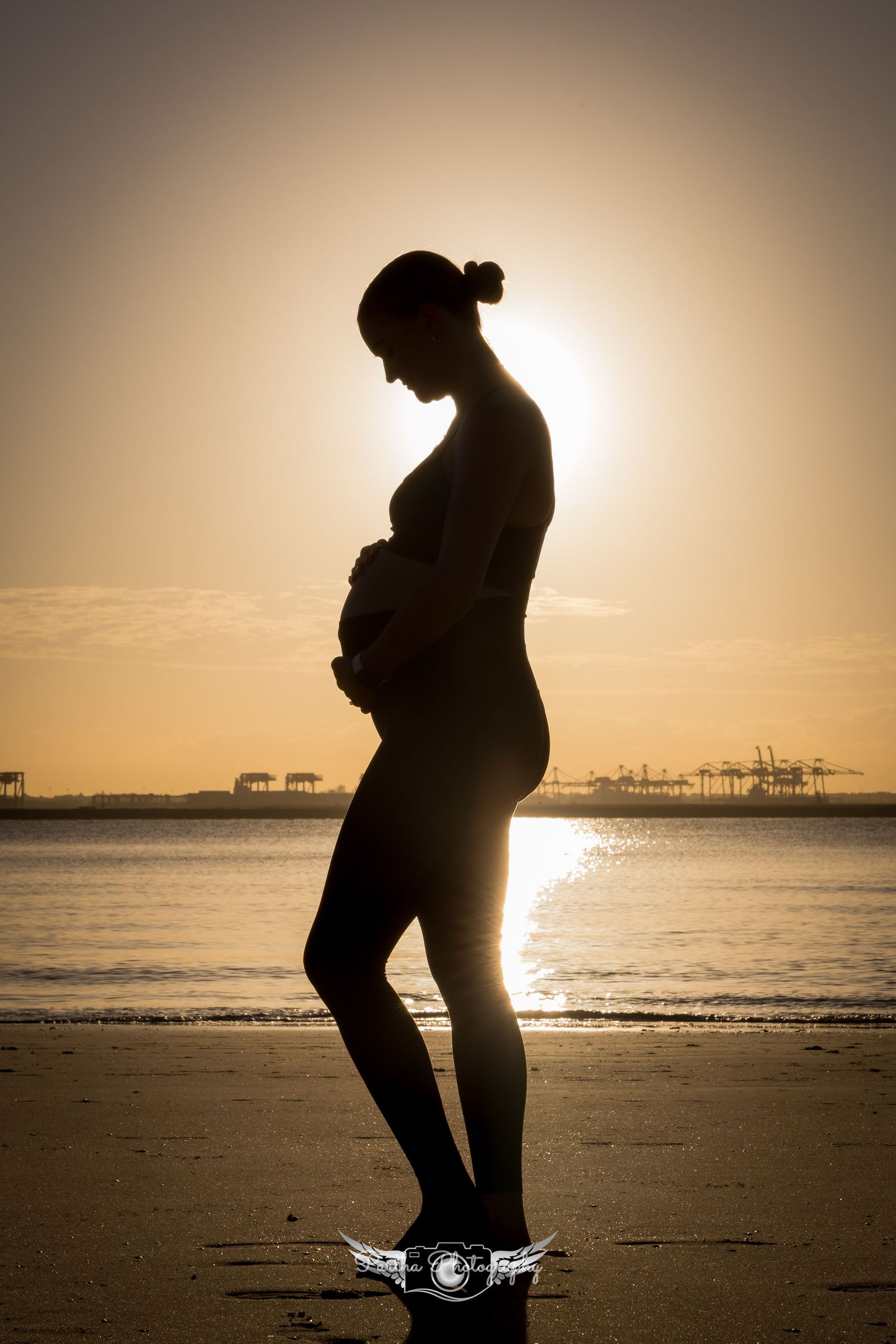 Silhouetted pregnant figure stands on beach at sunset with industrial cranes visible in distance creating dramatic profile.