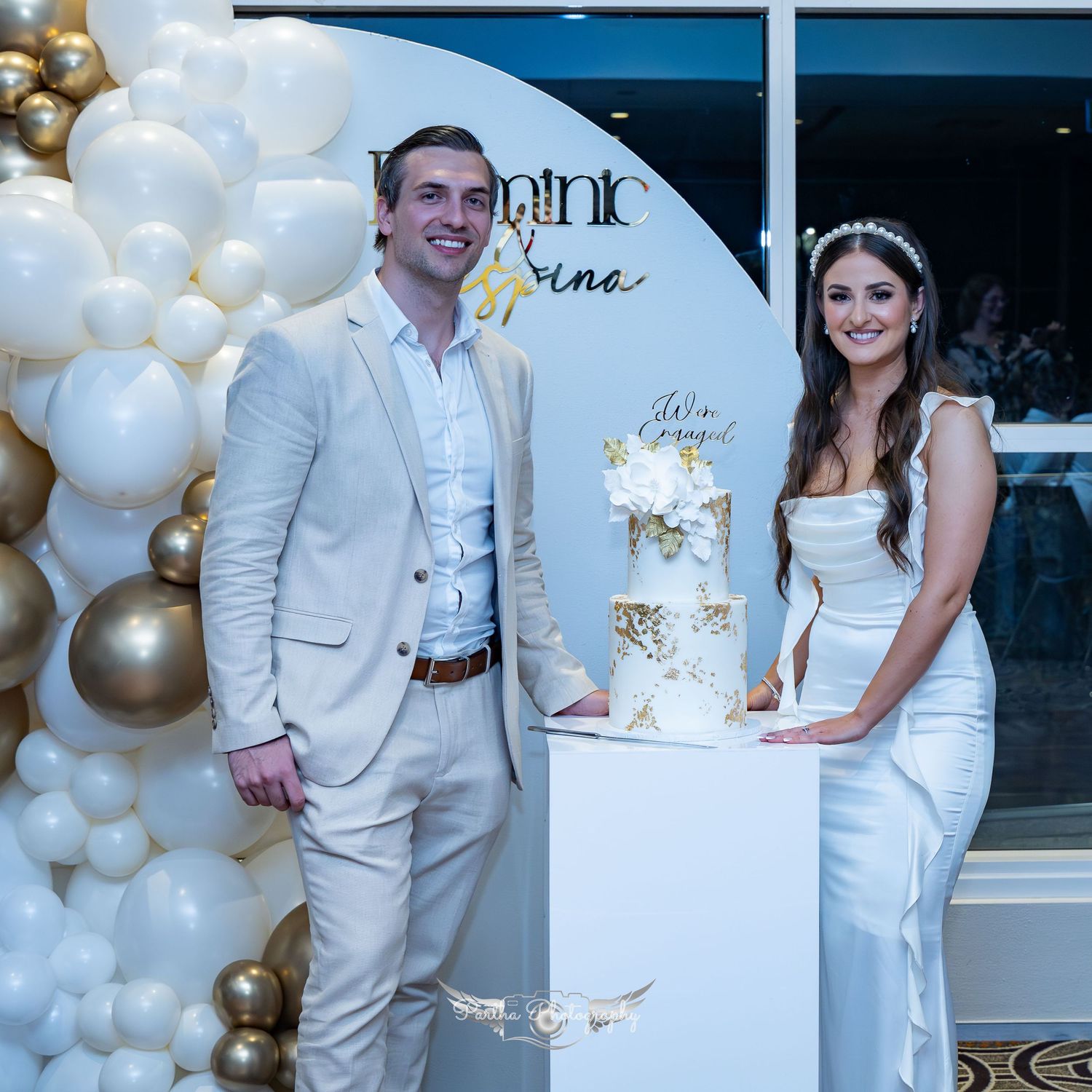 Couple stands beside elegant white and gold wedding cake with balloon decorations.
