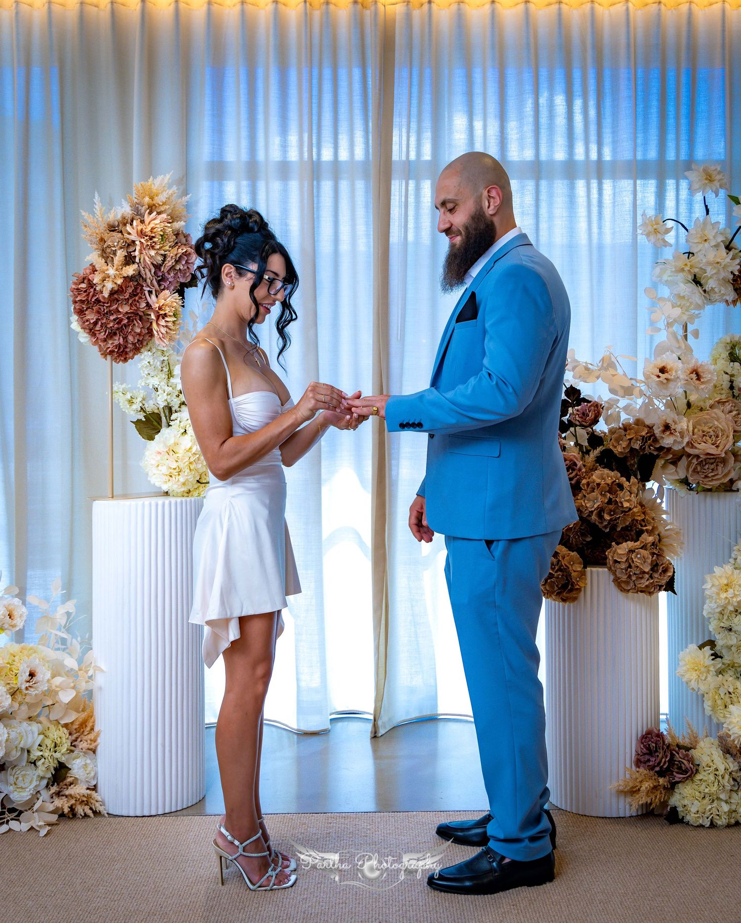 Wedding ceremony with couple exchanging rings surrounded by elegant floral arrangements and white pillars.