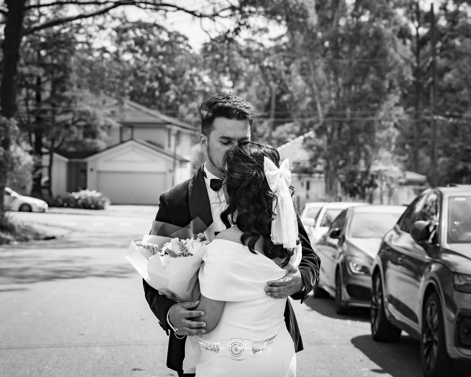A romantic black and white portrait of newlyweds embracing outdoors near parked cars.