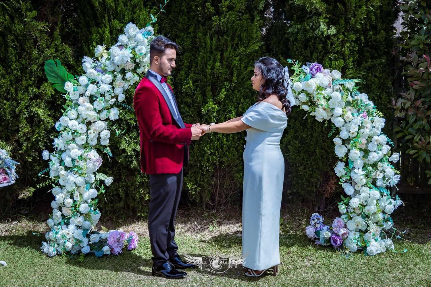 A couple exchanges vows under a beautiful floral arch with blue and white flowers in an outdoor garden.